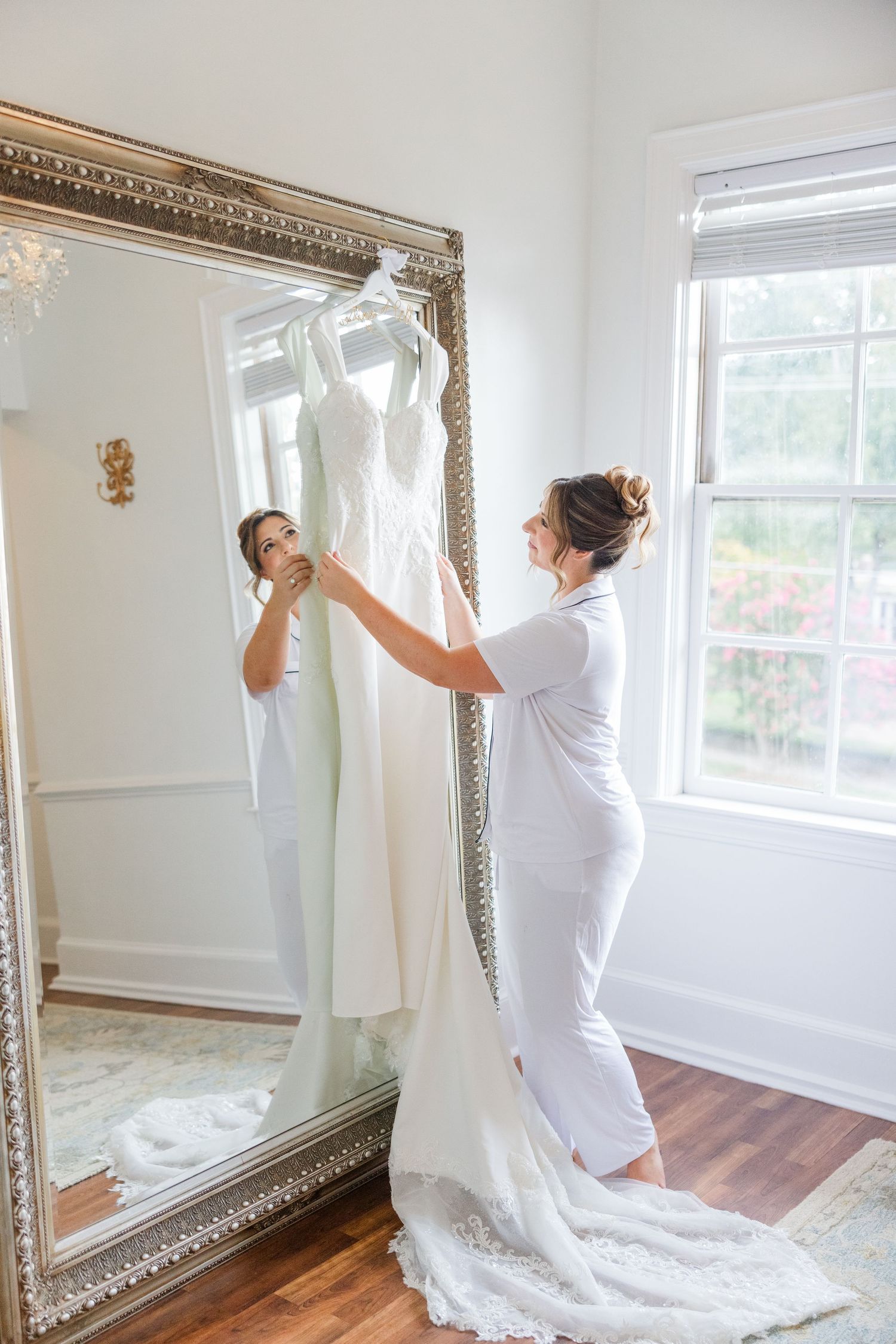 White wedding dress being hung on ornate mirror in sunlit room with hardwood floors.