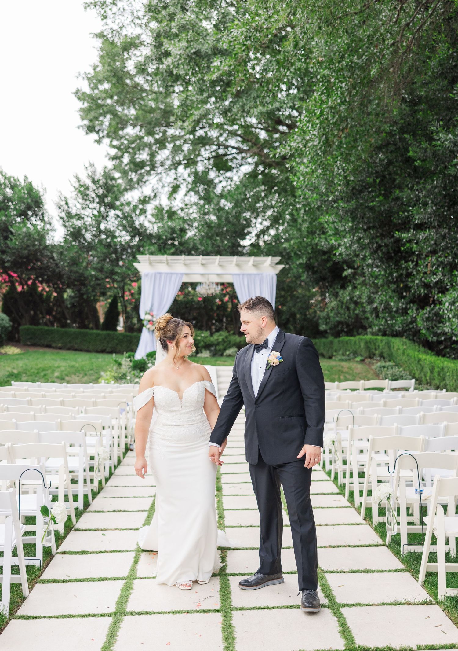 Couple shares intimate moments along white chairs set up for outdoor garden wedding ceremony.