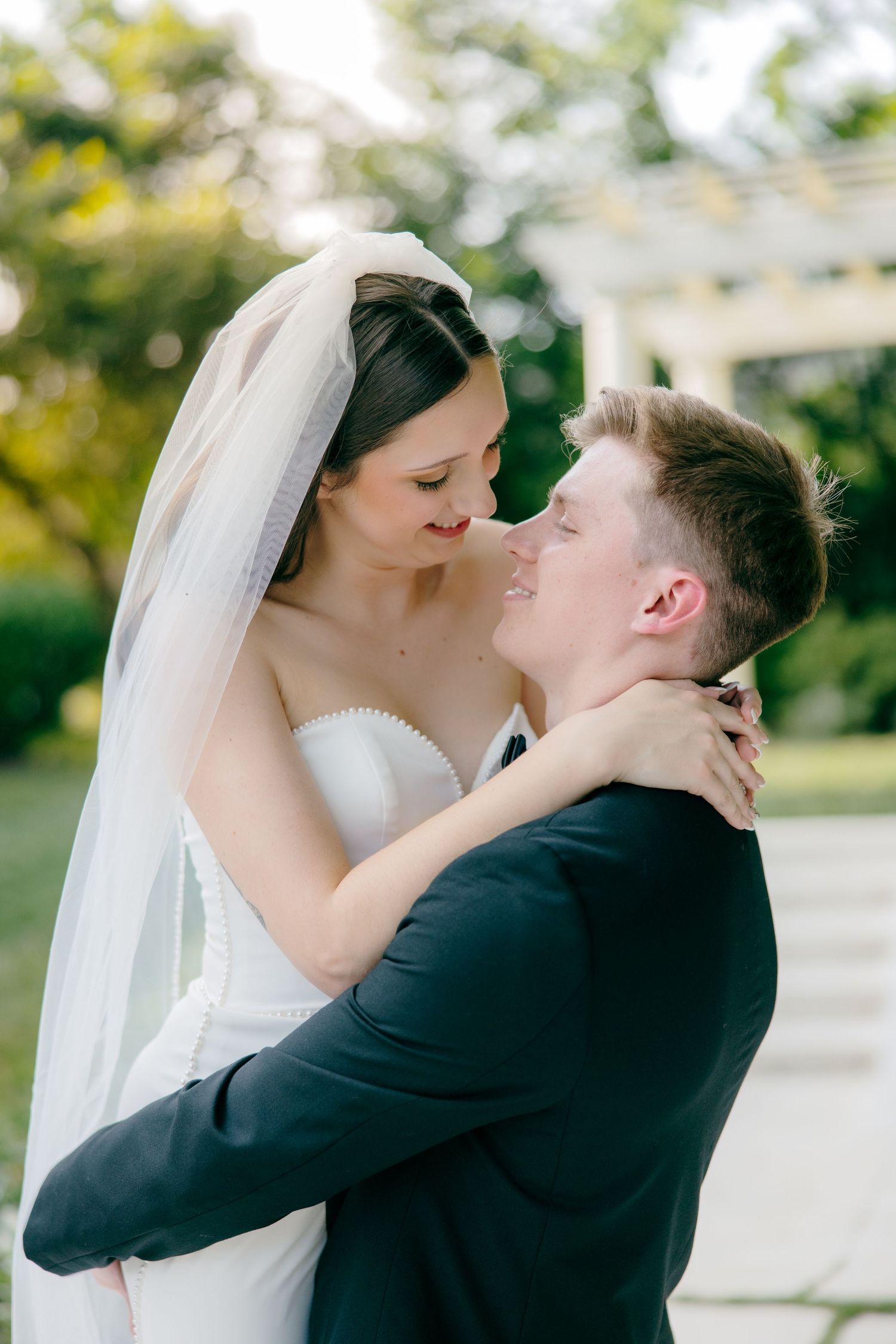 Intimate wedding moment captured in a sunlit garden with white pergola backdrop.