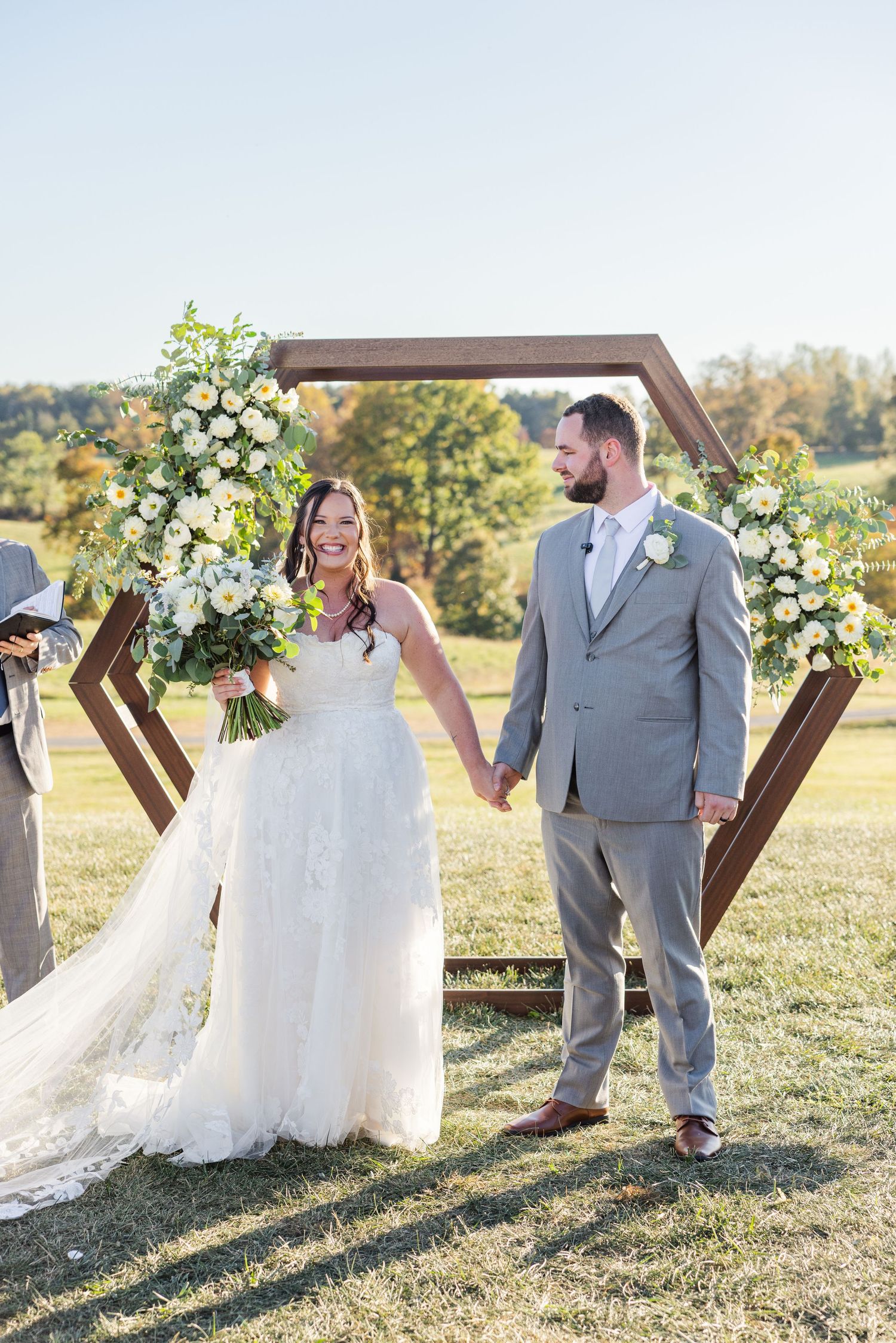Outdoor wedding ceremony setup with wooden cross arch and floral arrangements in a sunny field.
