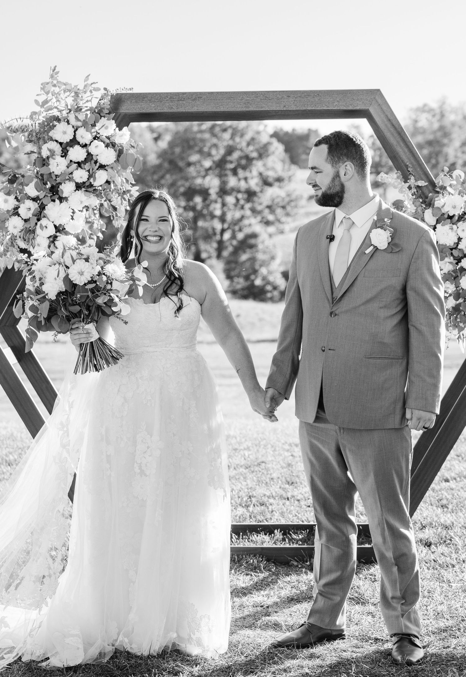 Newlyweds holding hands under geometric arch decorated with flowers in outdoor setting.