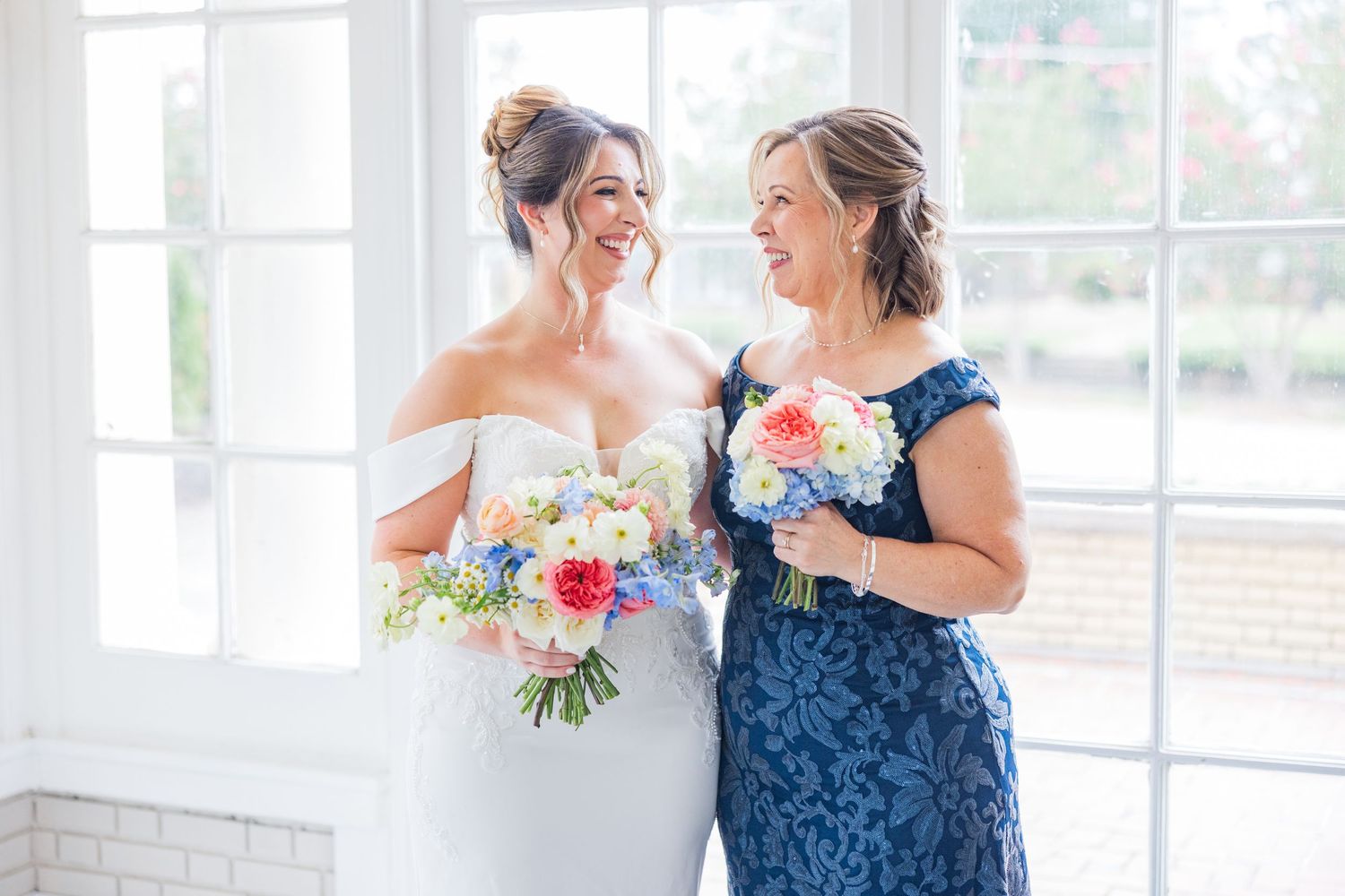 A bride in a white gown and a woman in navy blue share a joyful moment while holding floral bouquets.