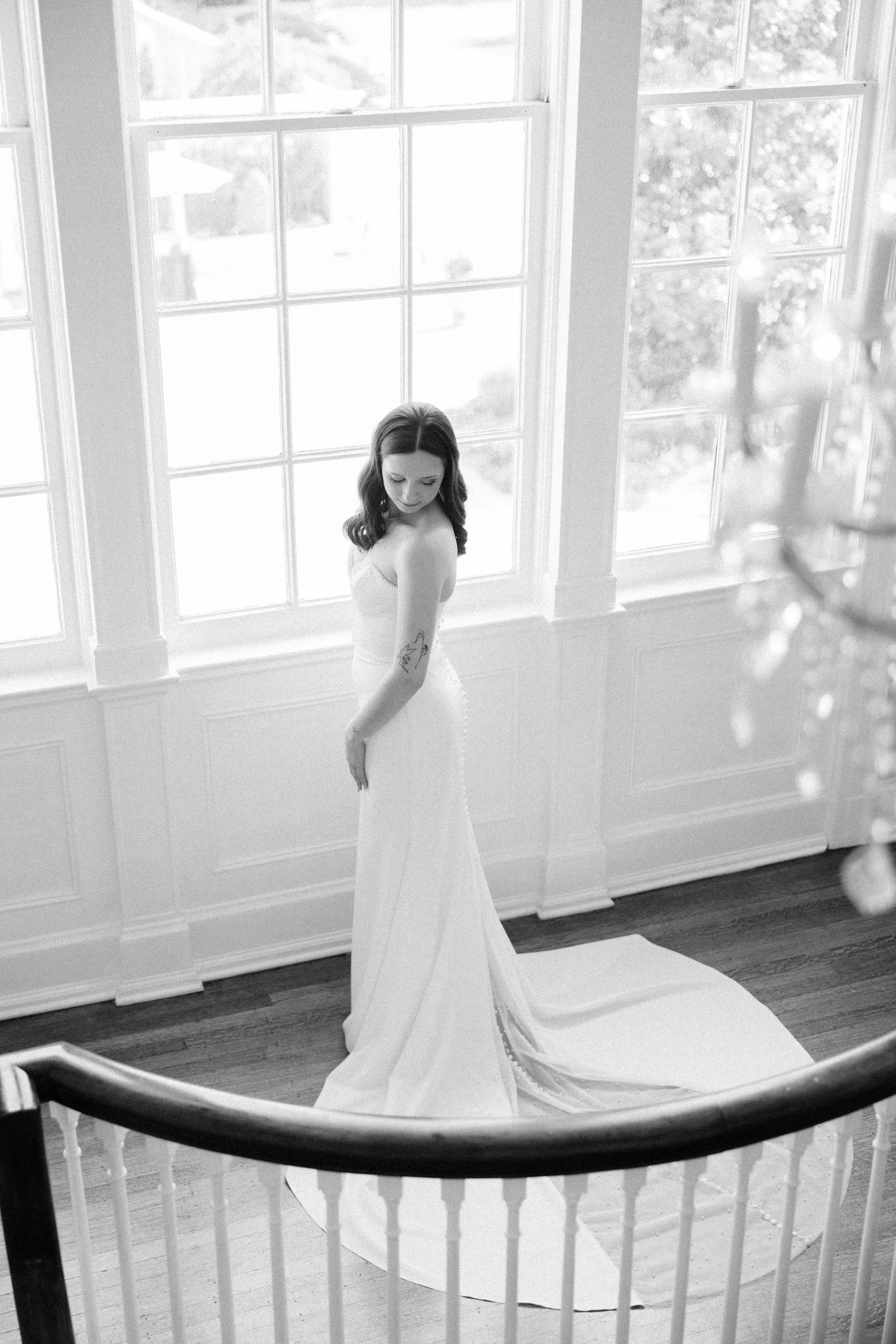 A black and white portrait of a bride in a fitted gown posing by a grand staircase window.
