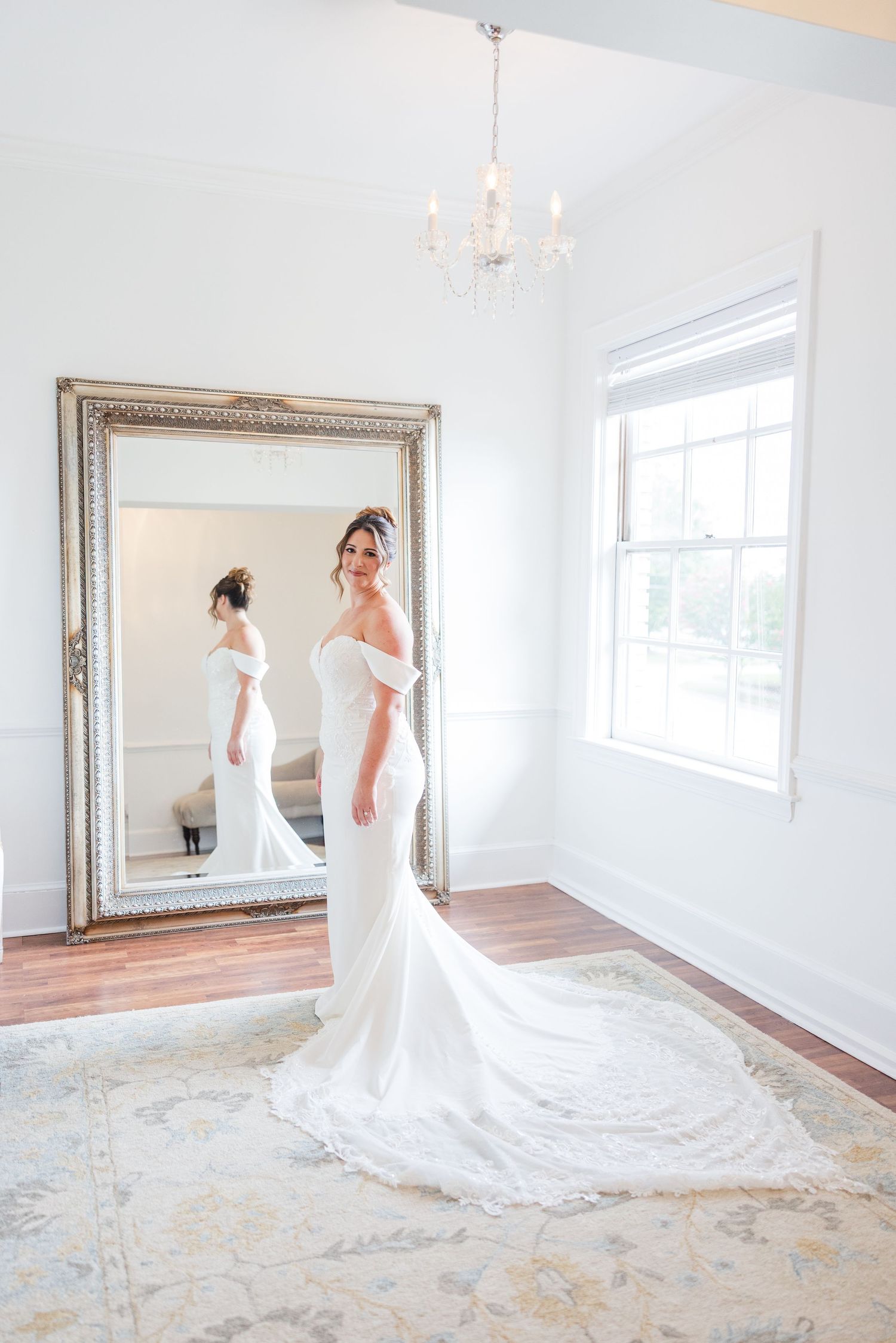 Elegant white wedding dress with long train displayed in a bright room with ornate mirror.