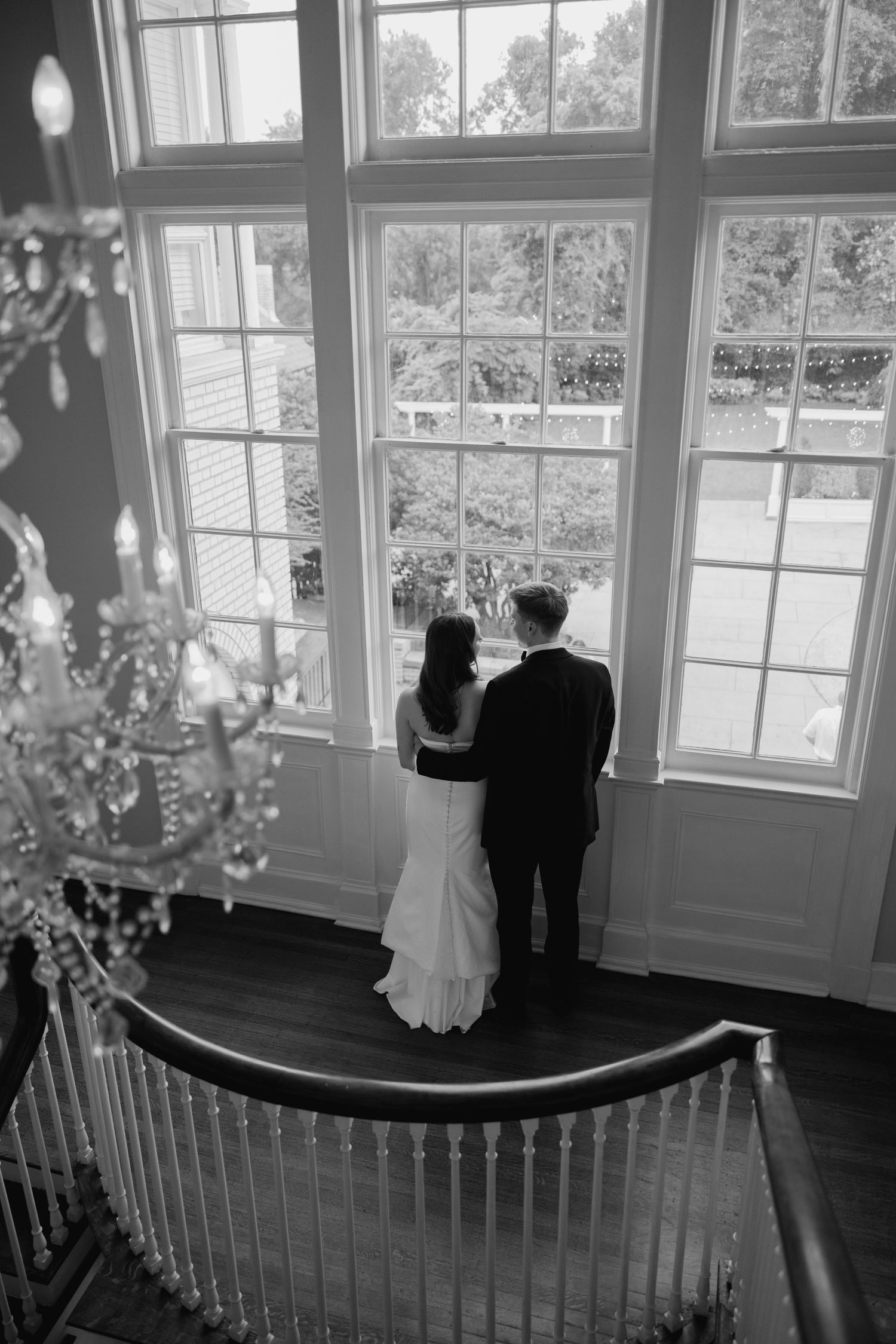 A black and white photo captures a couple dancing by large windows in an elegant room.