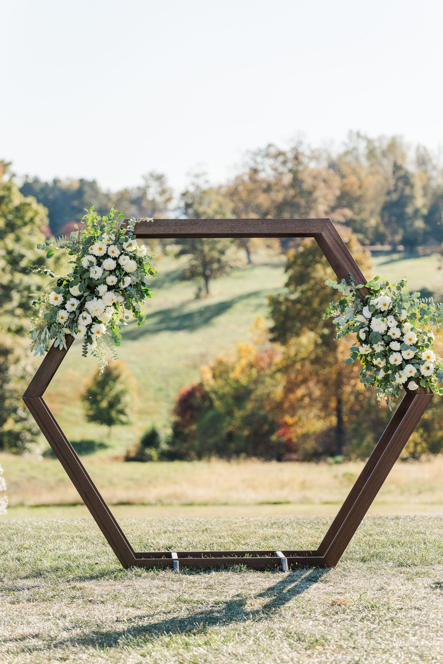Modern hexagonal wooden wedding arch decorated with white floral arrangements in an outdoor setting.