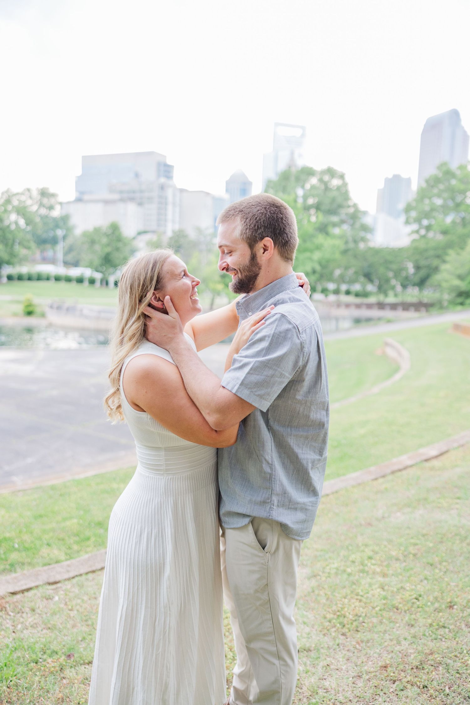 Romantic couple embraces on a grassy hill with city skyline in background during engagement photoshoot.