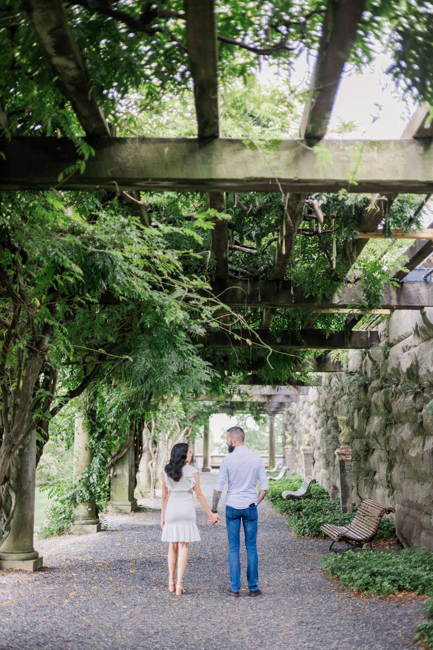 Couple strolls hand in hand under a rustic wooden pergola along a gravel path.