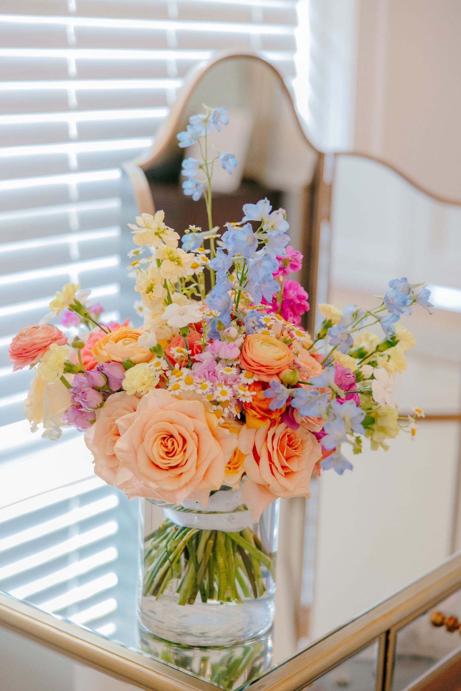 Vibrant floral arrangement with peach roses, blue delphinium, and pink blooms in a clear glass vase by a window.