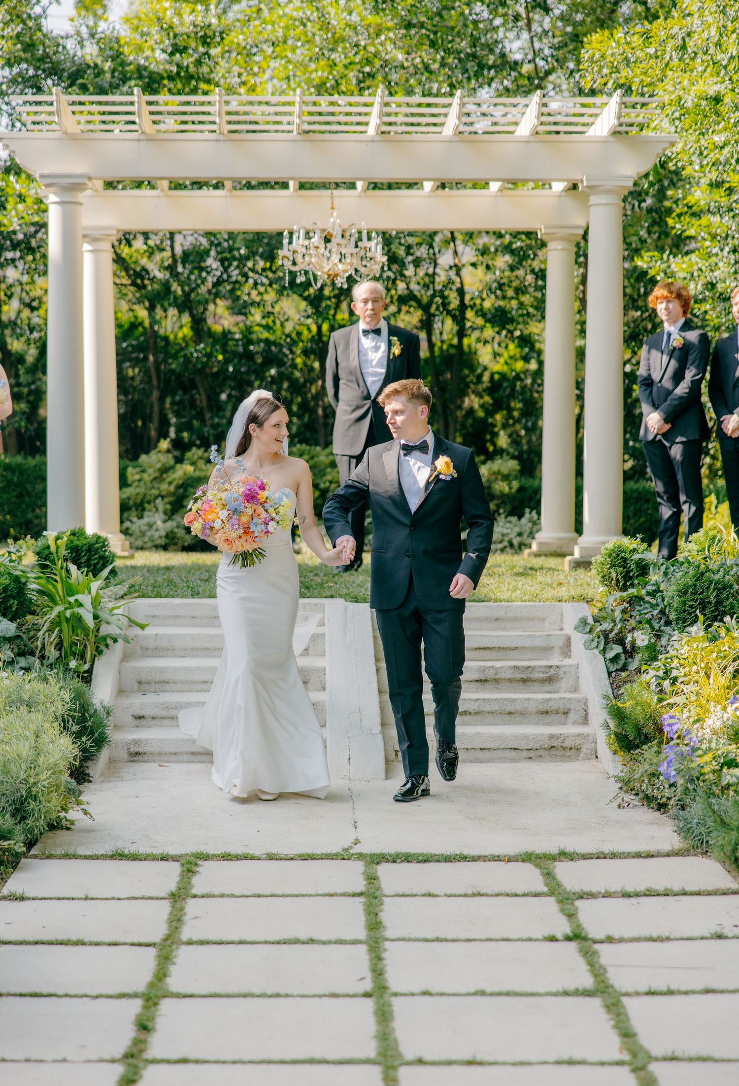 Wedding couple walking under white pergola in garden setting with stone pathway.