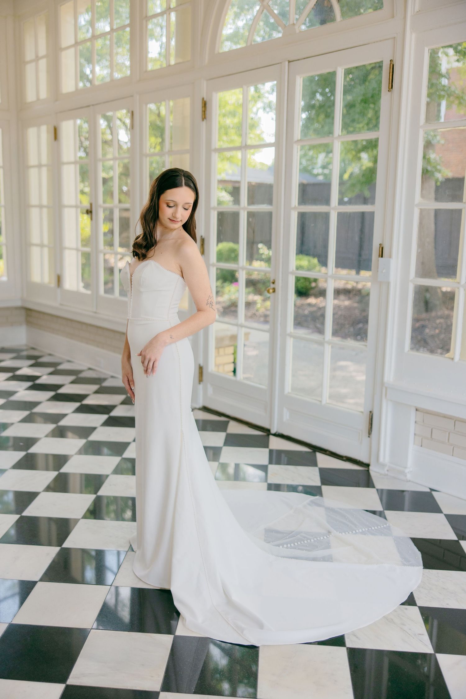 Bride in flowing white gown twirls gracefully on black and white checkered floor in sunlit room with large windows.