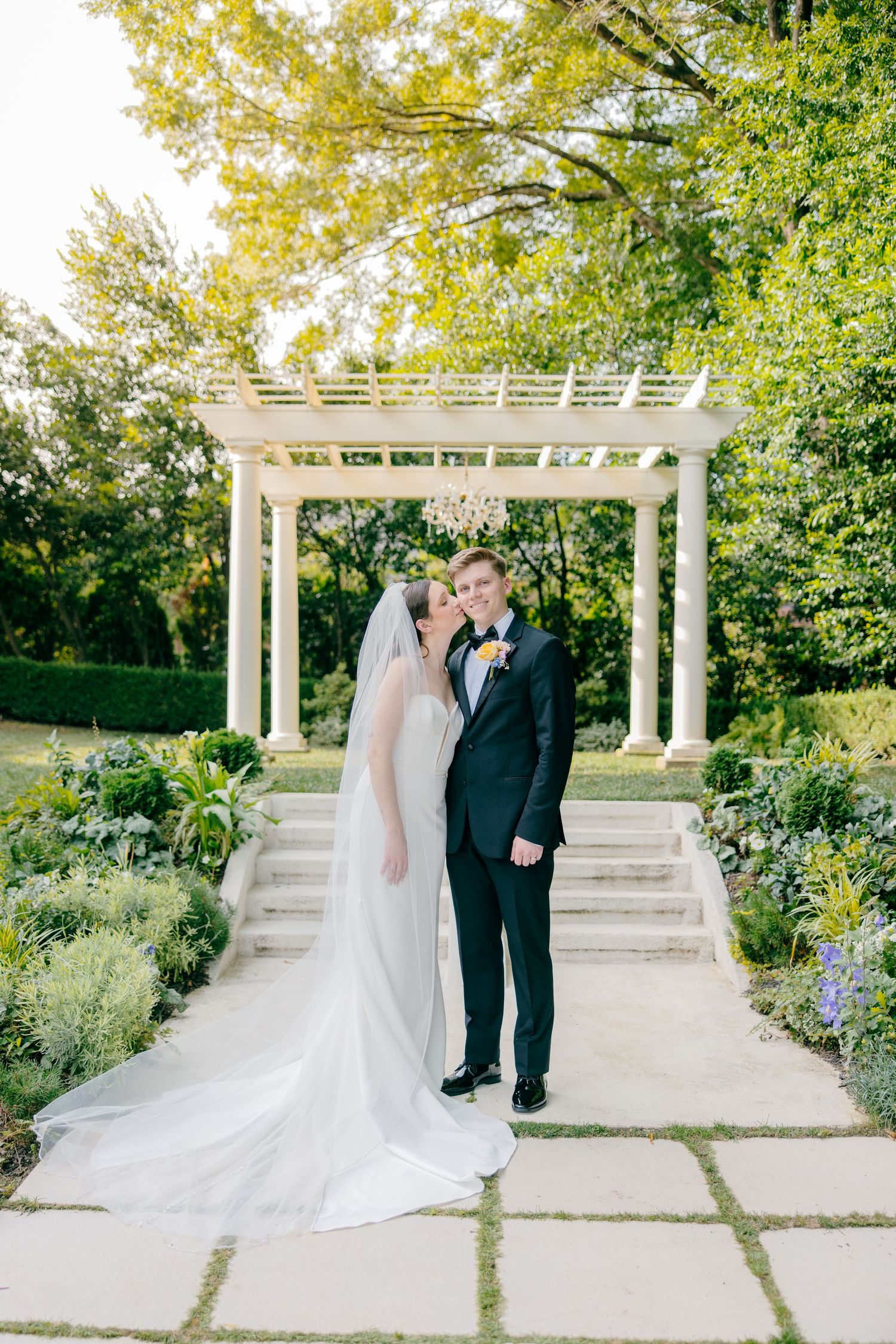 Newlyweds share a romantic moment under a white pergola in a garden setting.