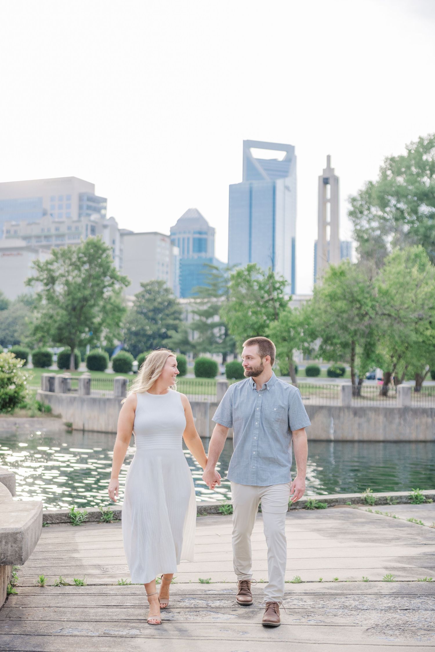 A couple walks along a waterfront path with city buildings and lily pads in the background.