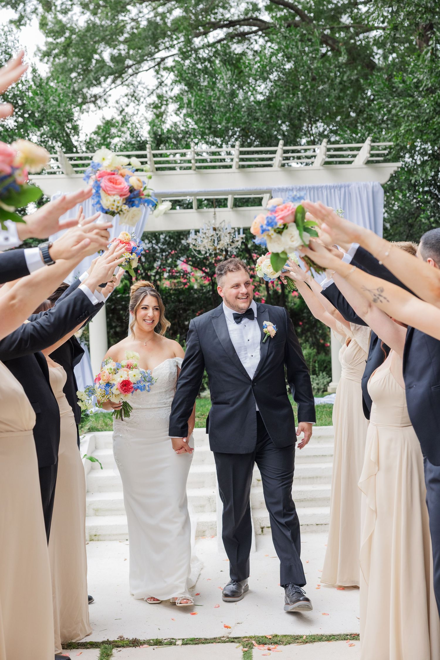 Wedding party creates arch with raised arms under white pergola outdoors.