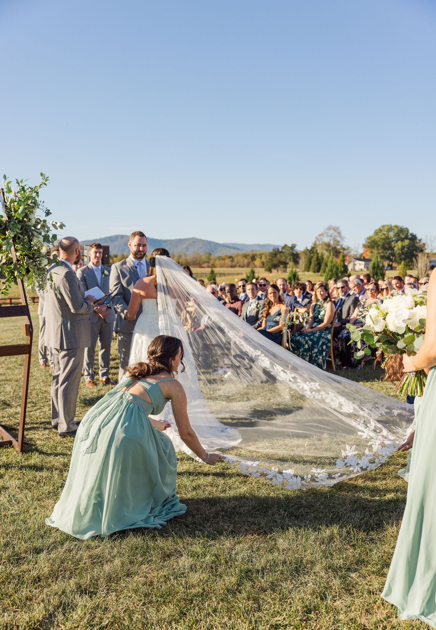 Wedding guests watch as a long veil flows in the wind across a grassy field at an outdoor ceremony.