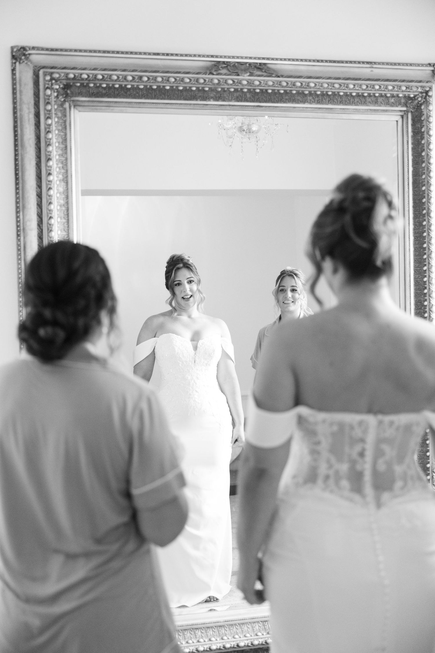 Group of women in formal attire stand before ornate mirrors in a bright room.