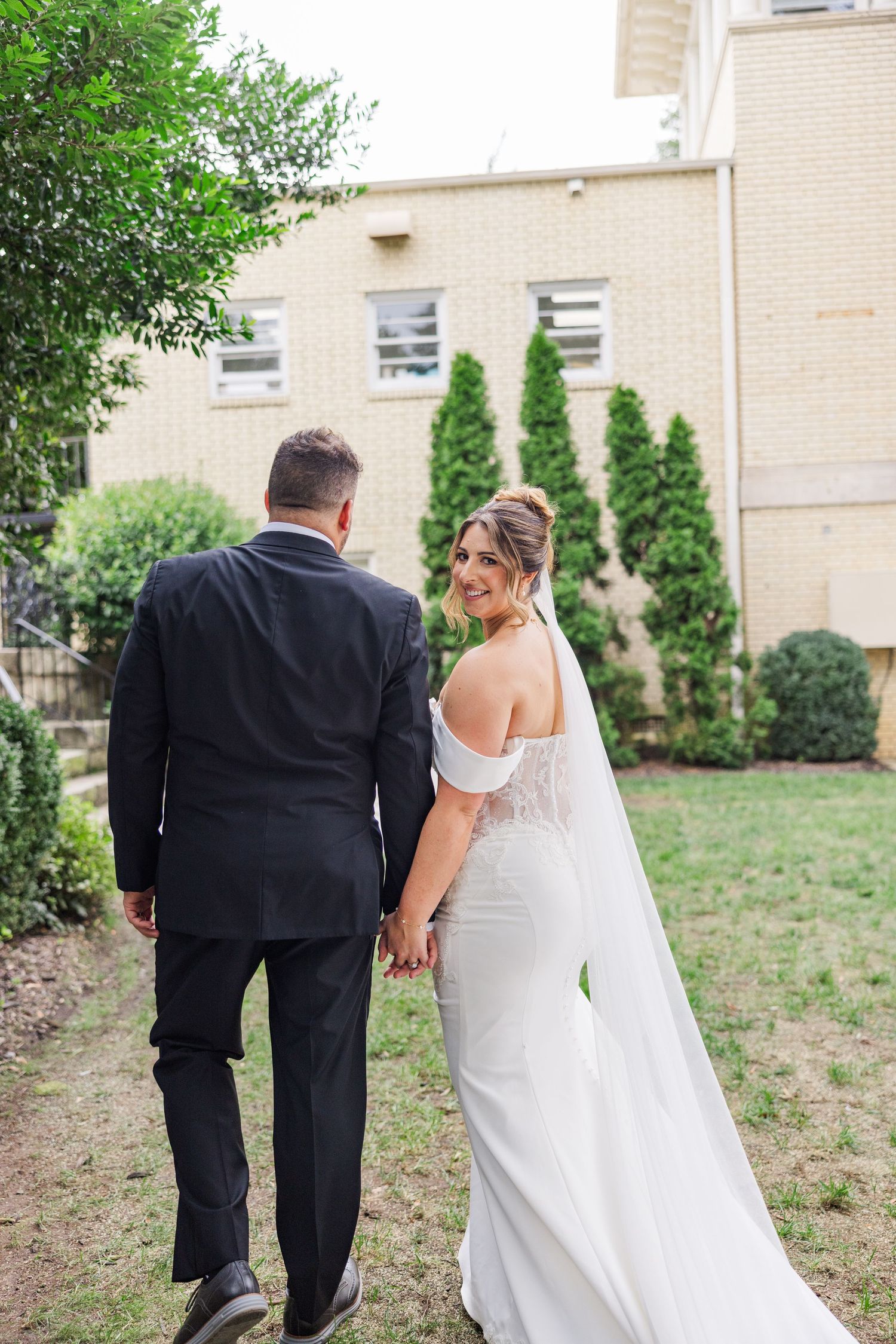 Couple walking hand in hand through a garden path beside a brick building.