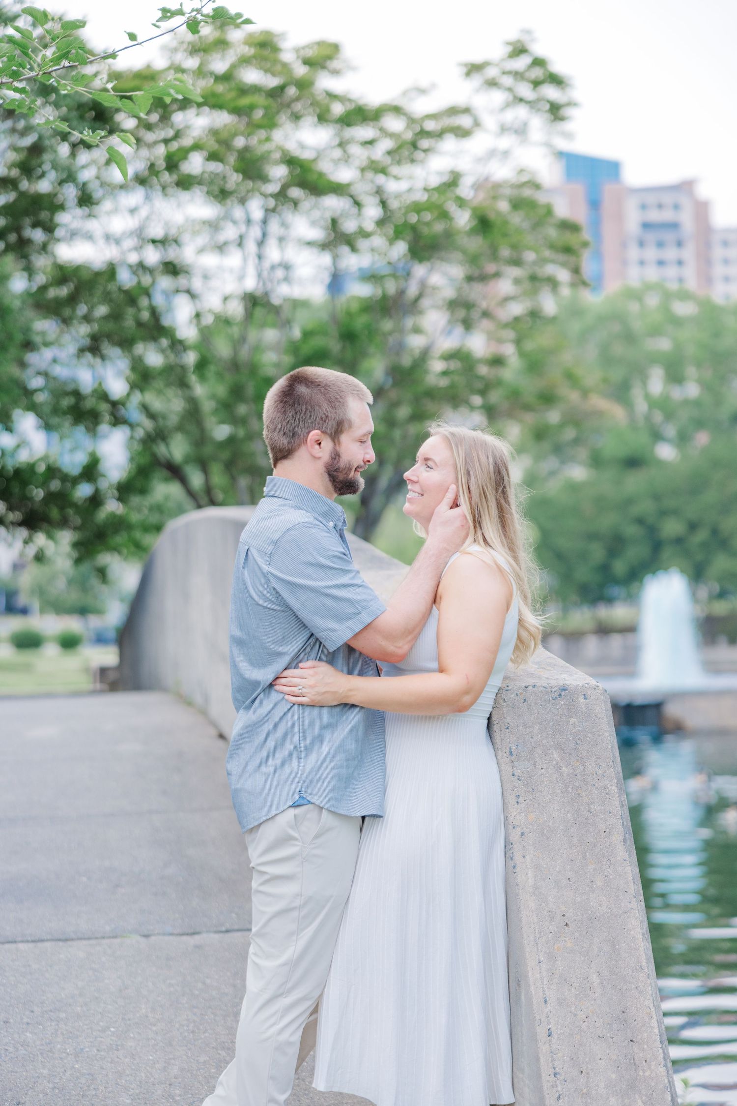 A couple shares tender moments by a waterfront with trees and buildings in the background.