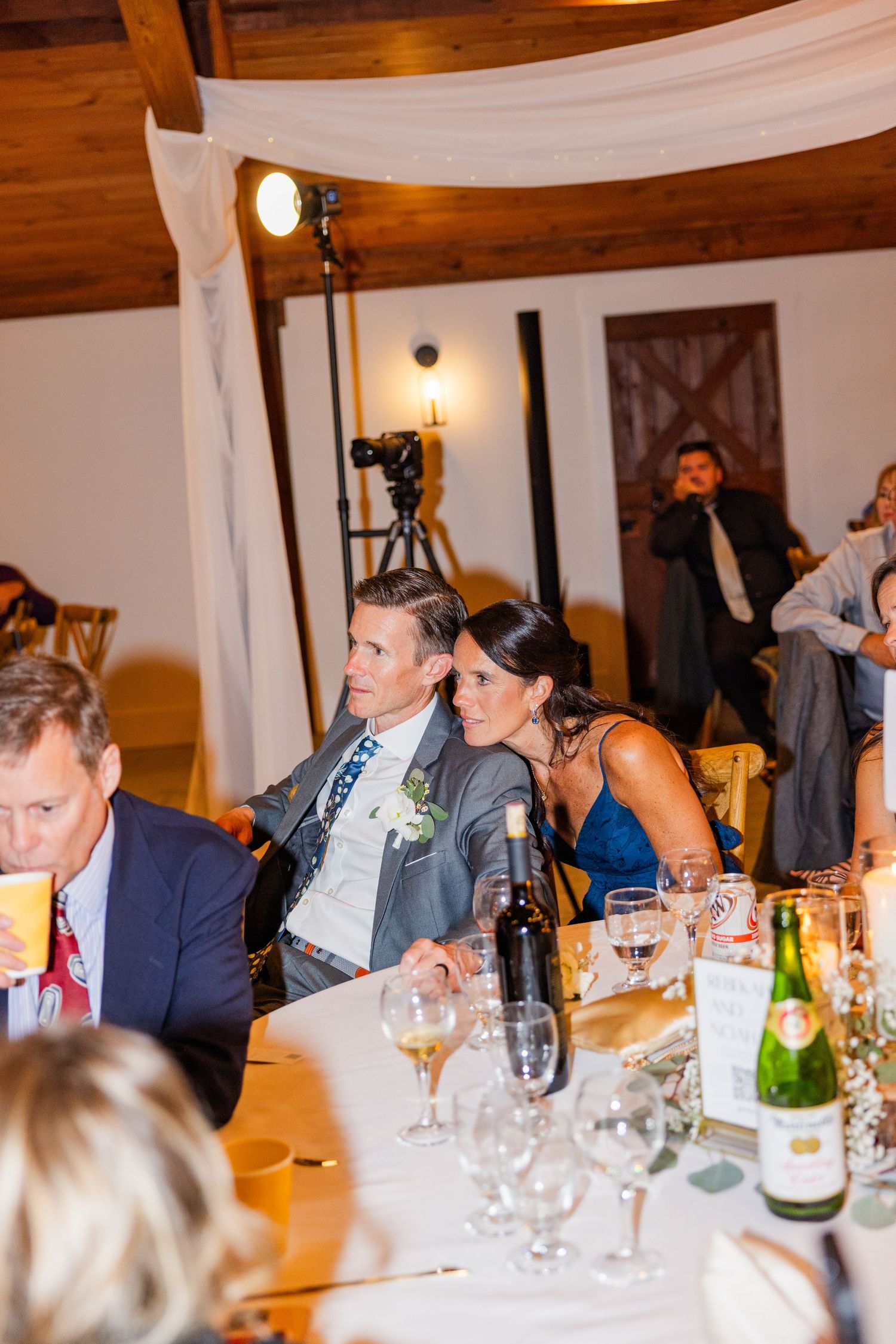 Wedding guests seated at a long dinner table with wine glasses and white draping decorations overhead.