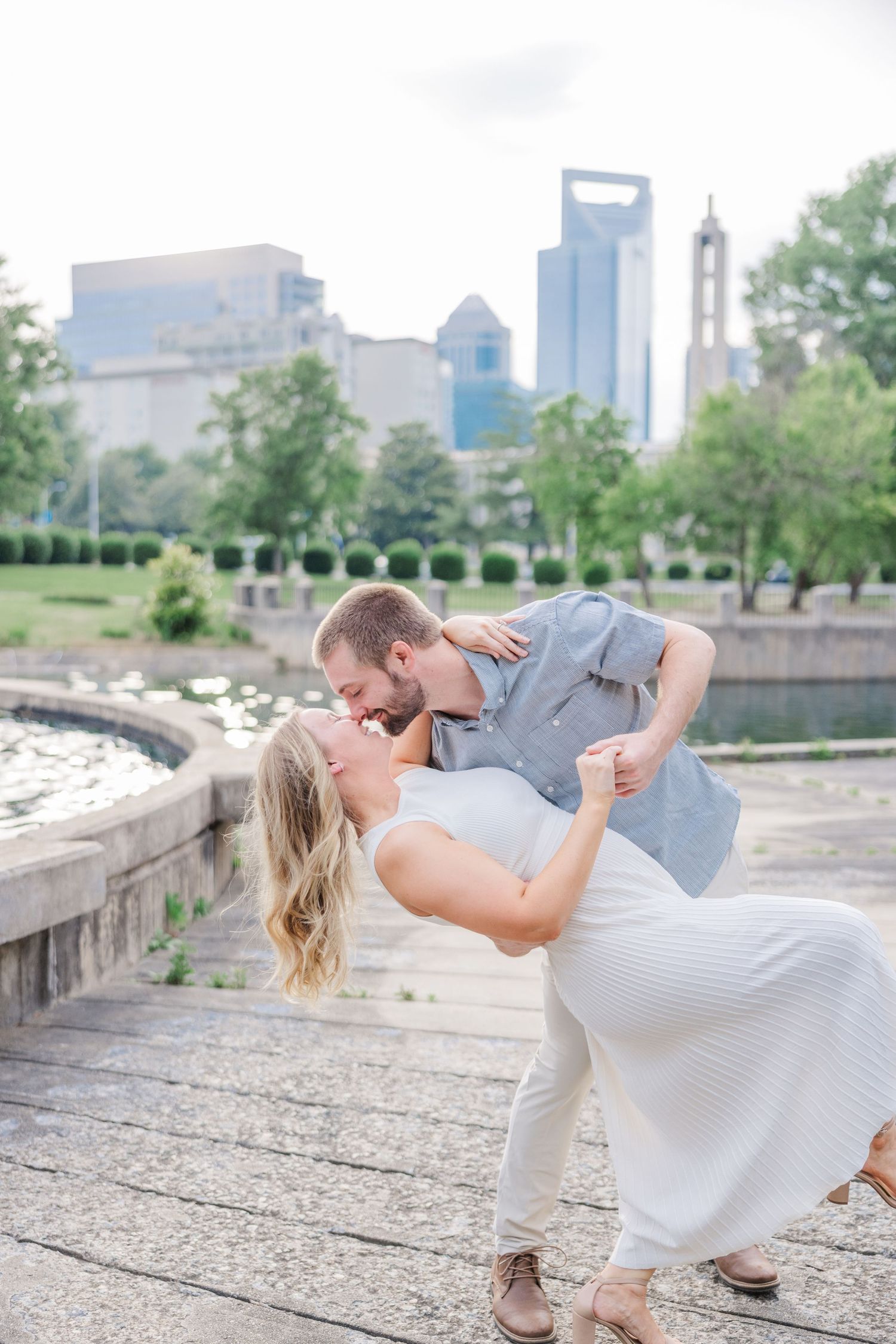 A romantic couple shares a dip kiss pose against the backdrop of the Charlotte skyline in a park setting.