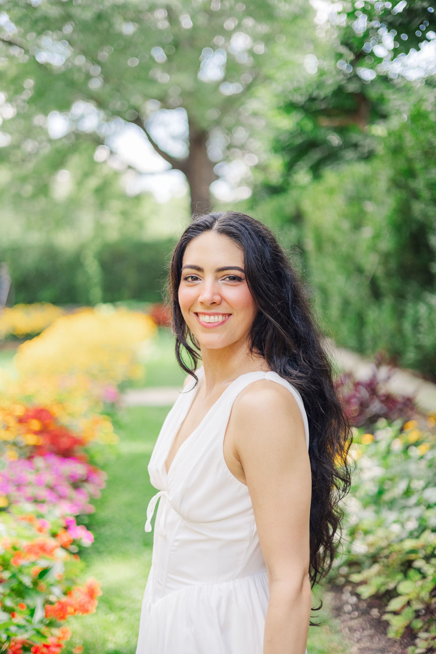 A woman in a white dress poses in a garden filled with colorful summer blooms.