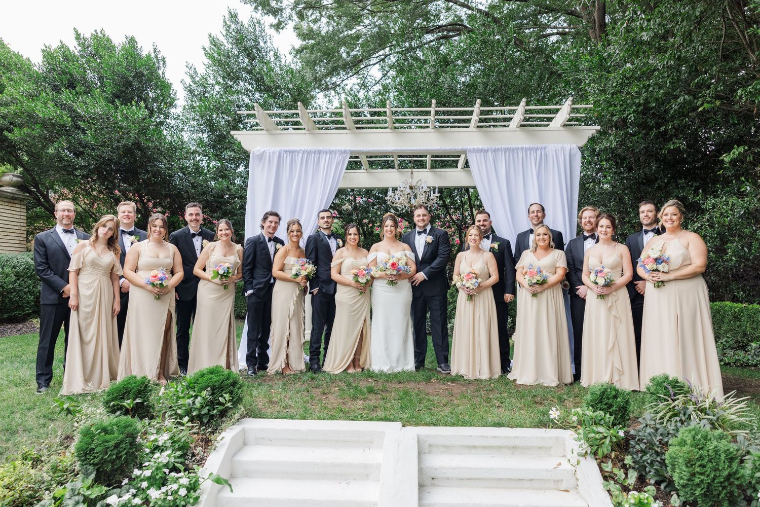 A wedding party poses outdoors in coordinated champagne-colored formal attire on a summer day.