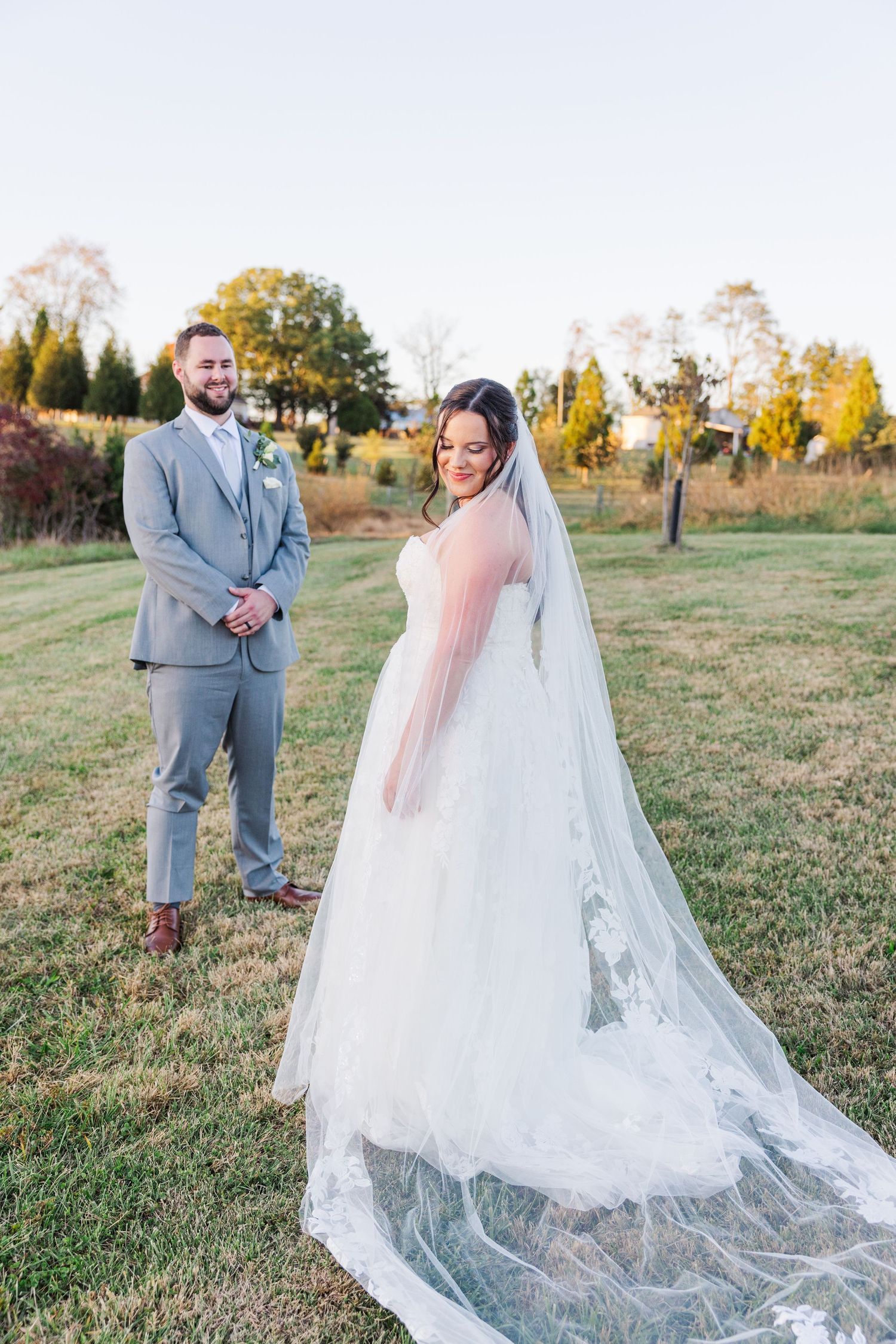 Wedding couple walks together across a grassy hill with the bride's lace veil trailing behind.