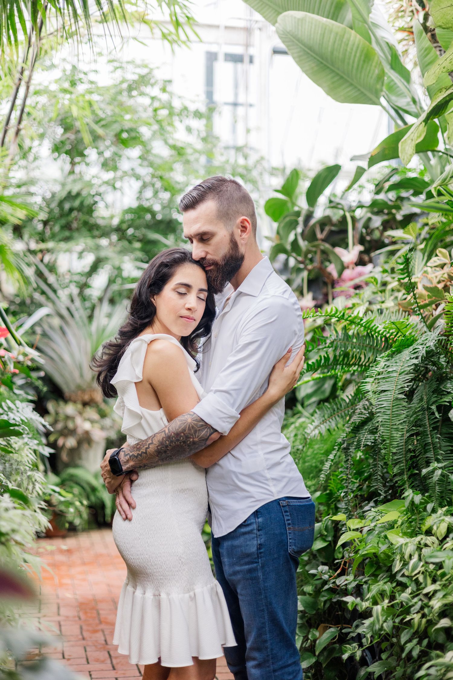 A romantic couple embraces in a lush greenhouse setting surrounded by tropical plants and greenery.