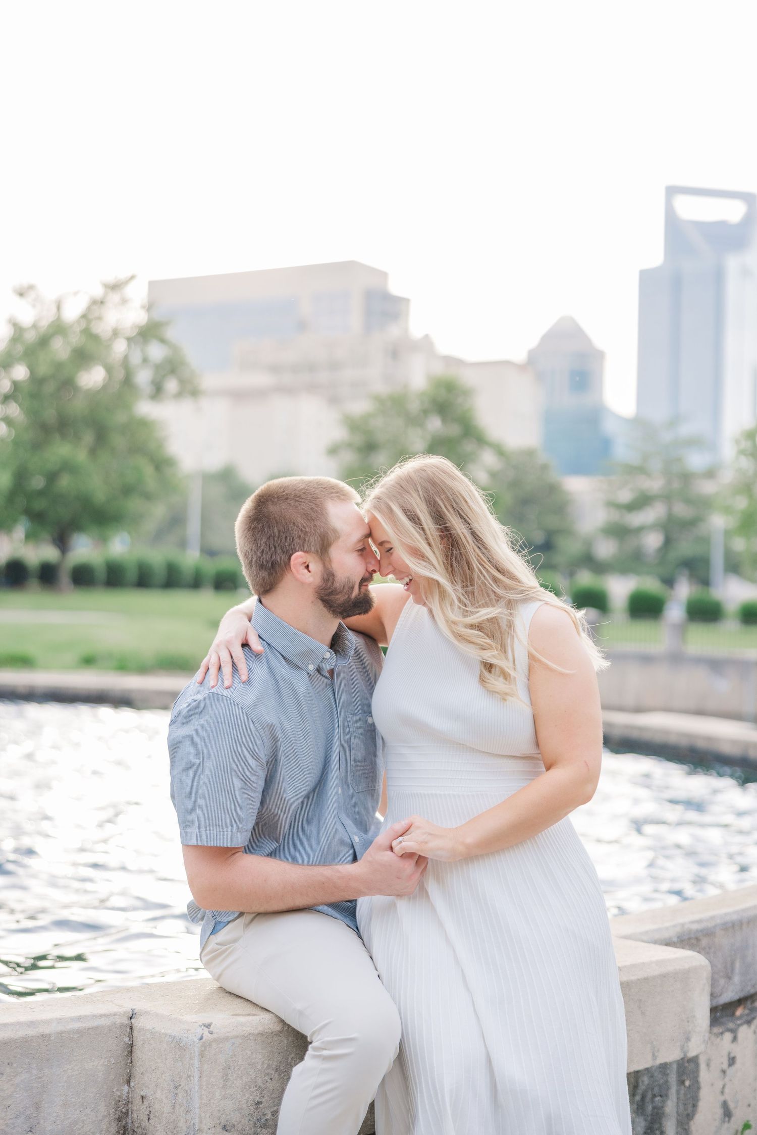 Couple shares intimate moments on stone wall by waterfront with urban skyline visible.
