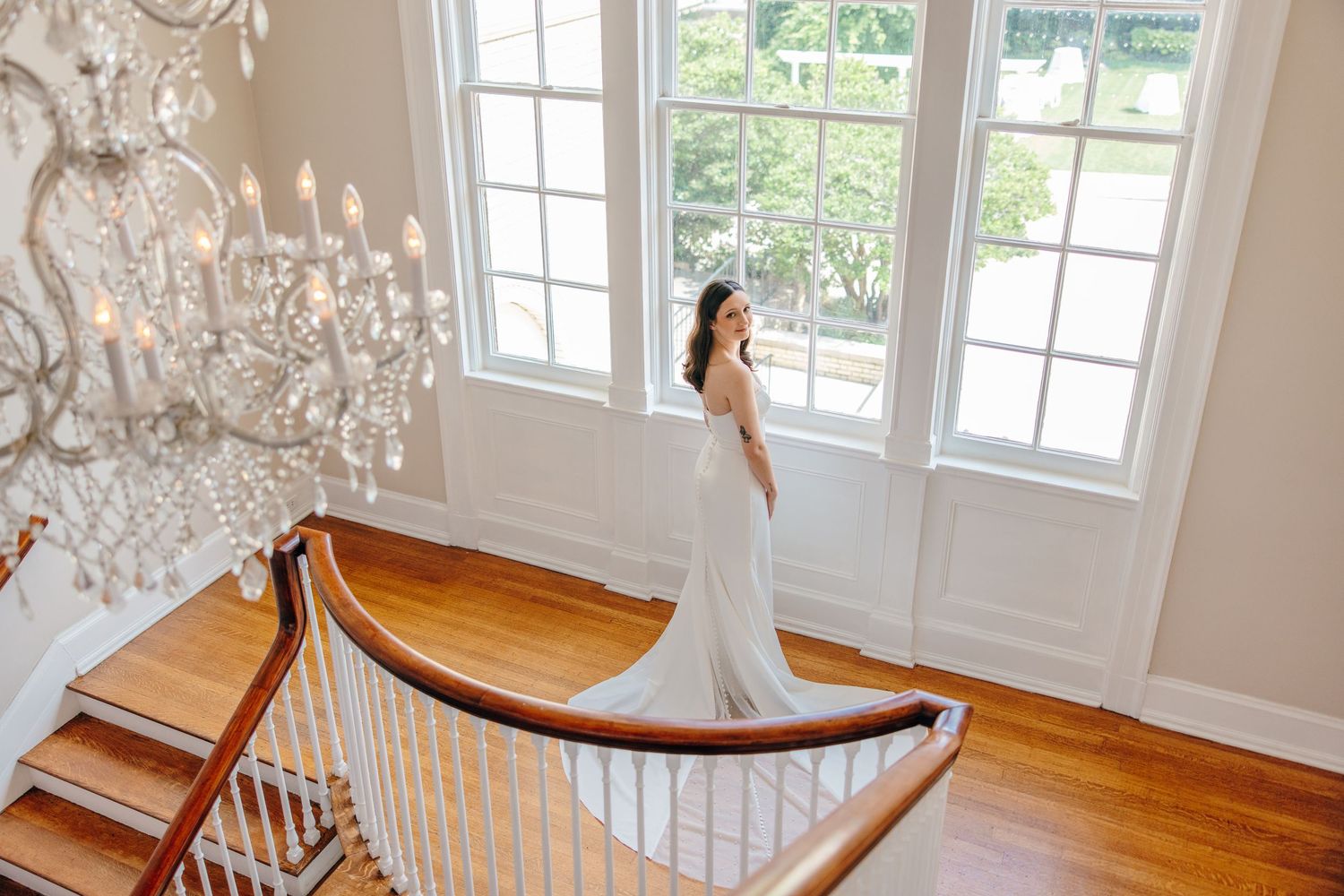 Elegant white wedding dress photographed in a bright historic mansion with crystal chandelier and curved wooden staircase.
