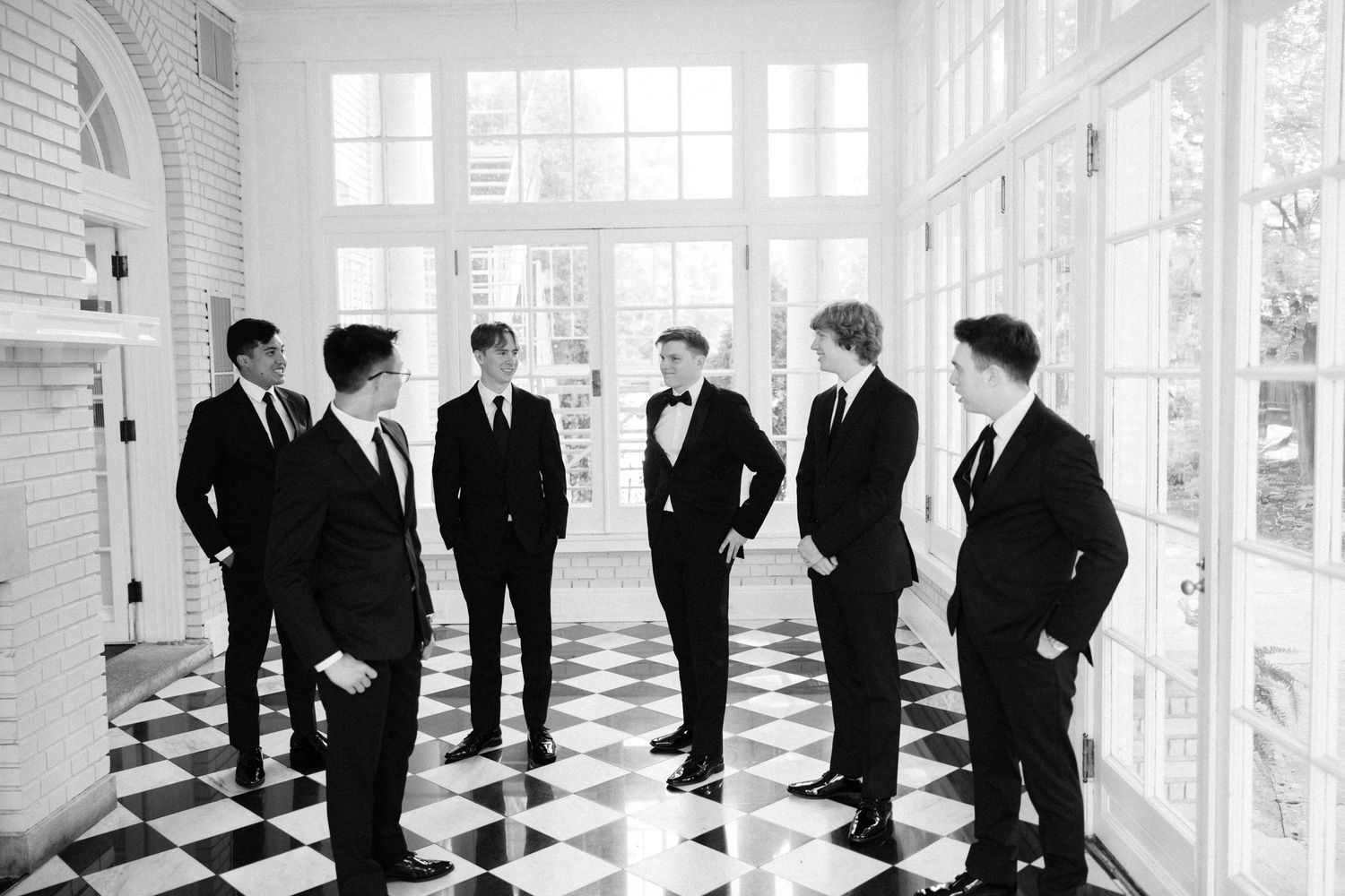 A group of groomsmen in black suits gathered in a hallway with checkered floor tiles.