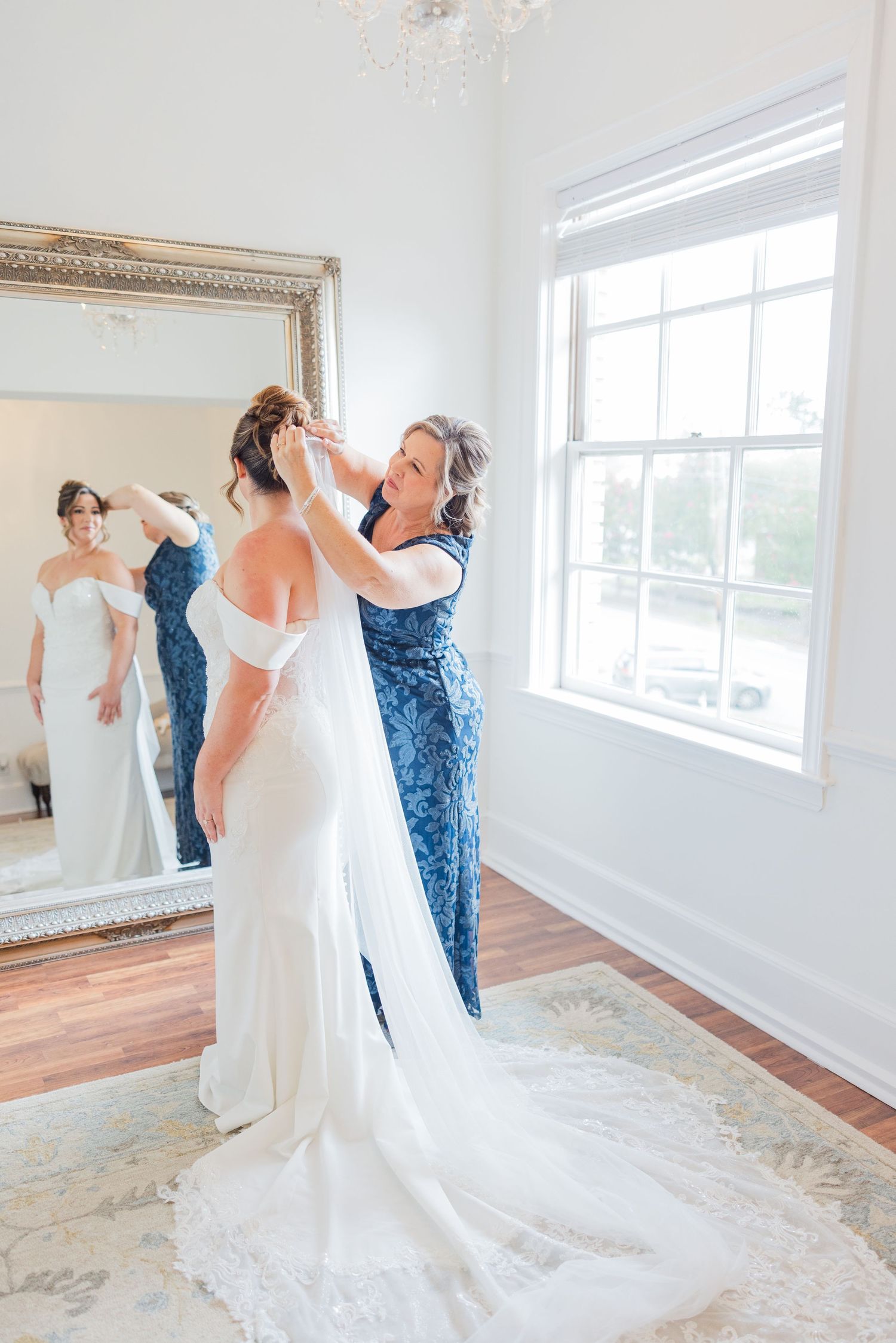 Bride in flowing white gown poses in front of large mirrors in sunlit room.