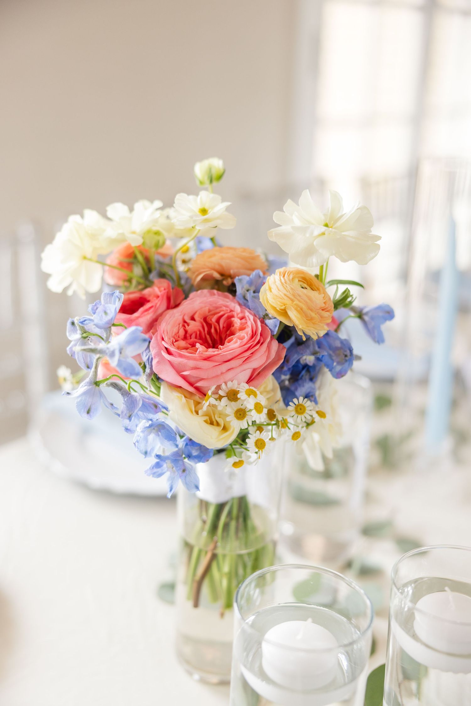 Delicate floral centerpieces with pink roses, blue delphiniums, and white flowers in glass vases on a light table.