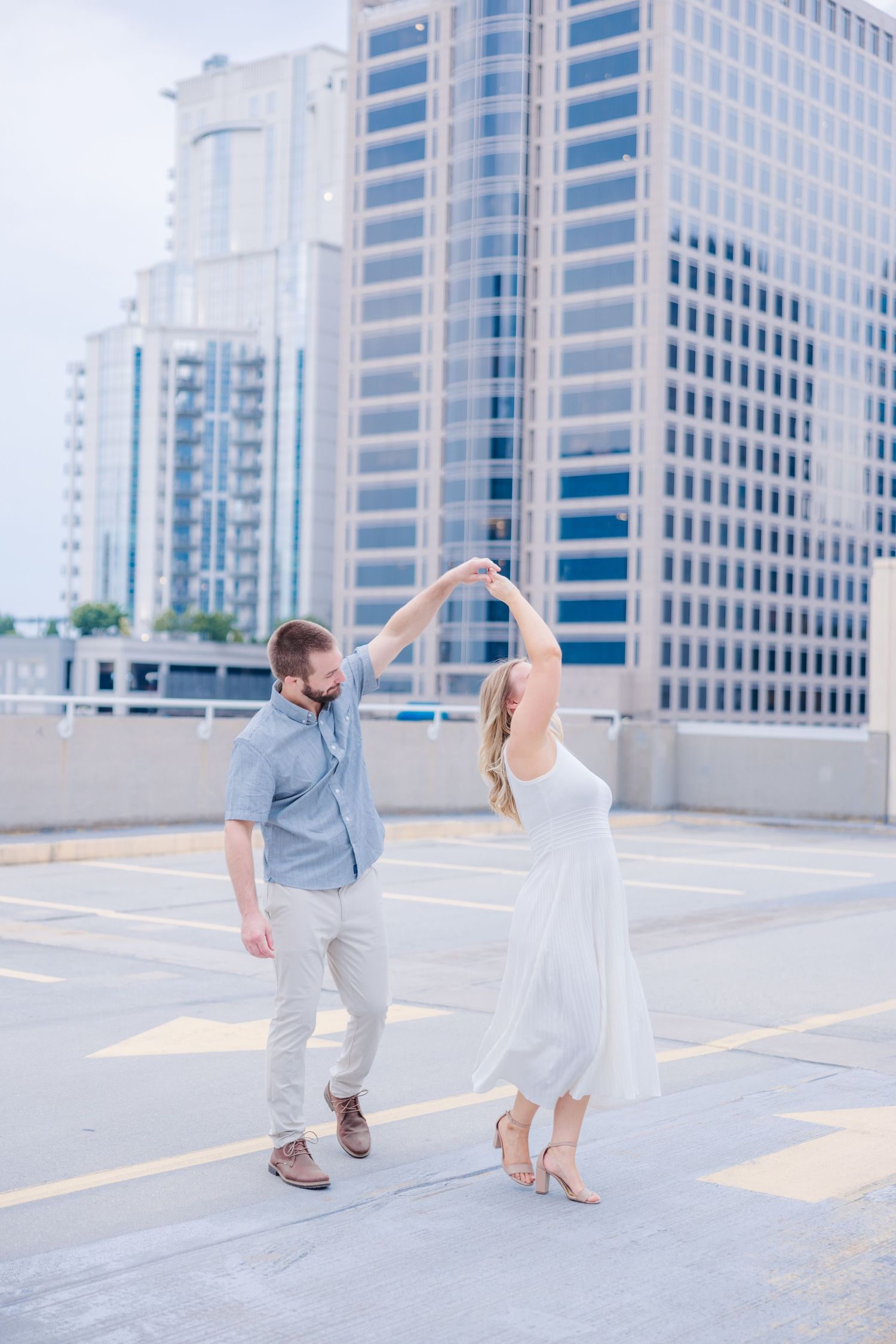 A couple dances playfully in an urban rooftop parking area with office buildings in the background.