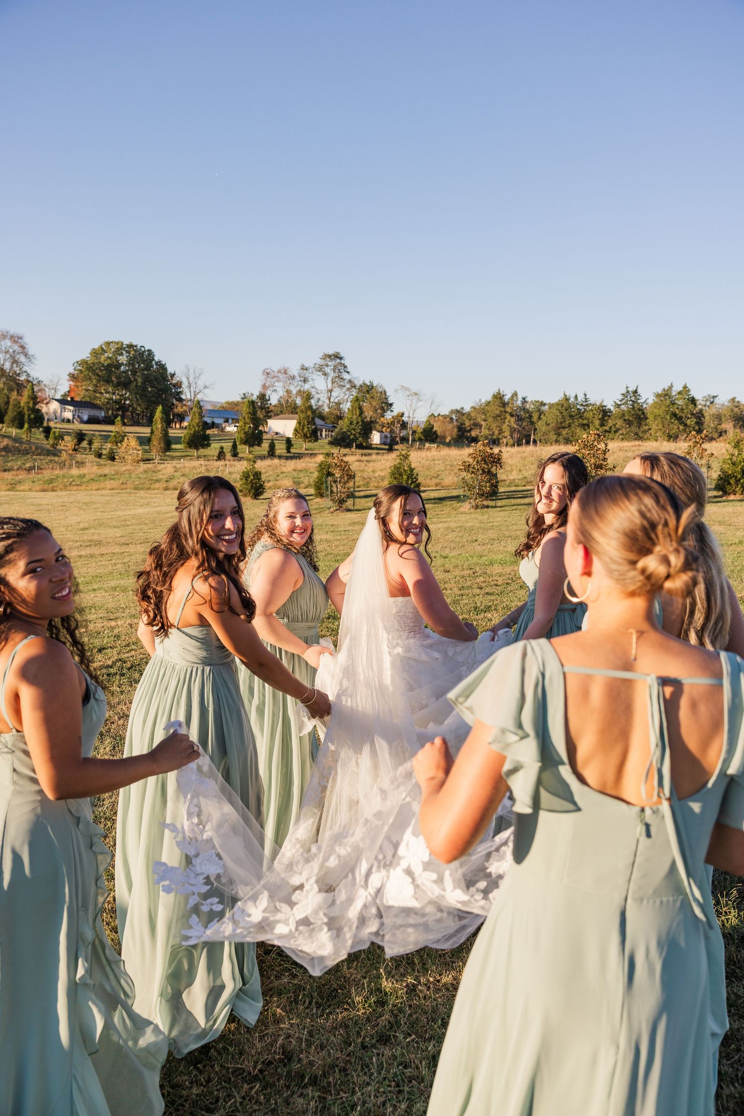 Bridesmaids in sage green dresses gather around the bride in an outdoor setting during sunset.