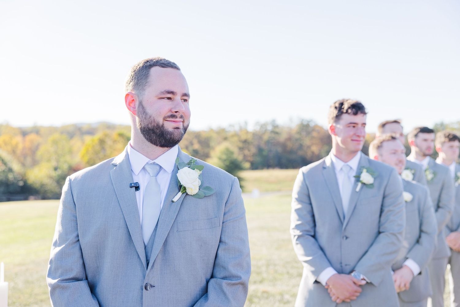 Groomsmen in light gray suits stand together outdoors on a sunny day during a wedding celebration.