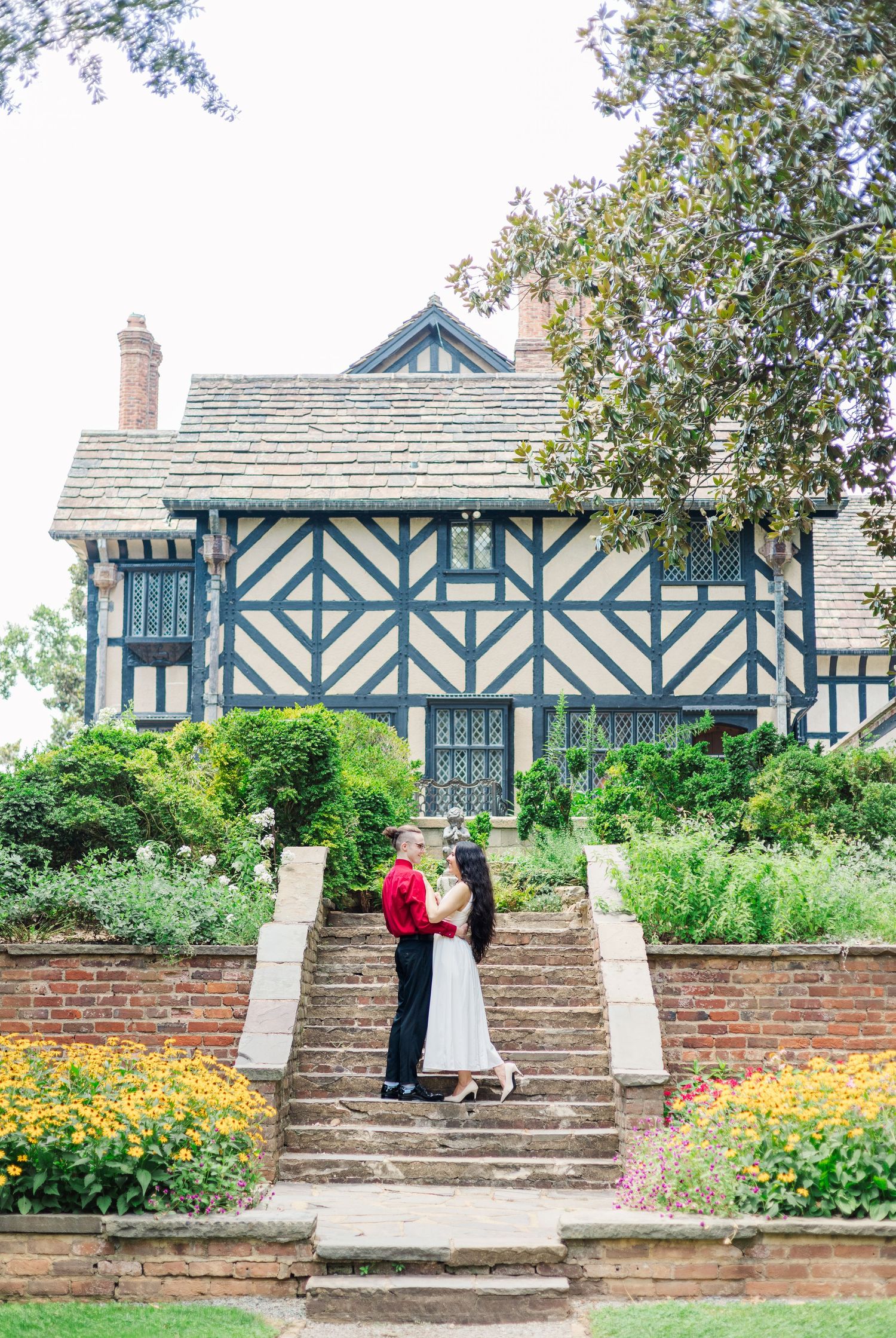 A couple stands on stone steps in front of a Tudor-style house with blue timber framing and blooming yellow flowers.