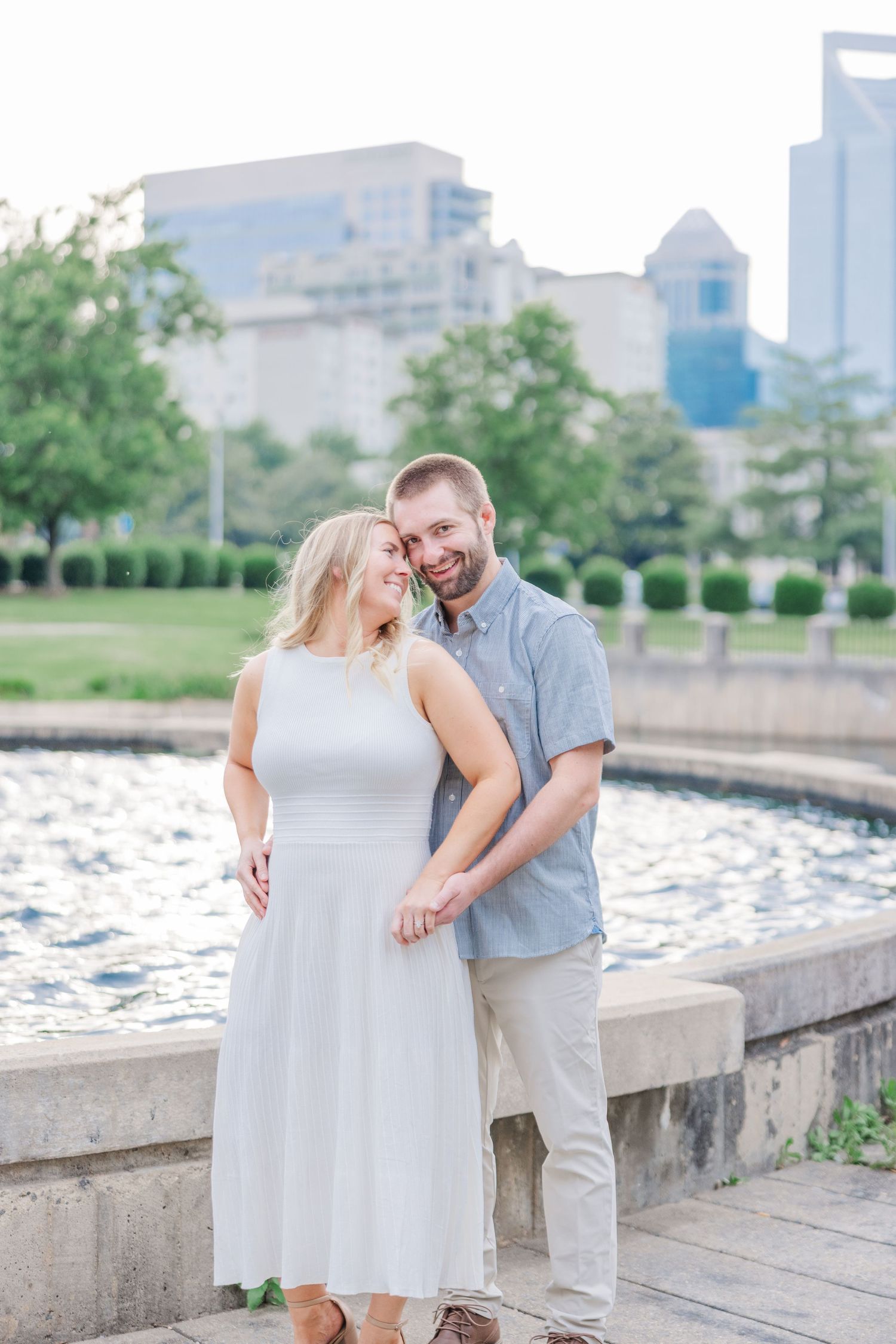 Romantic couple embraces along a waterfront promenade with city buildings in the background.