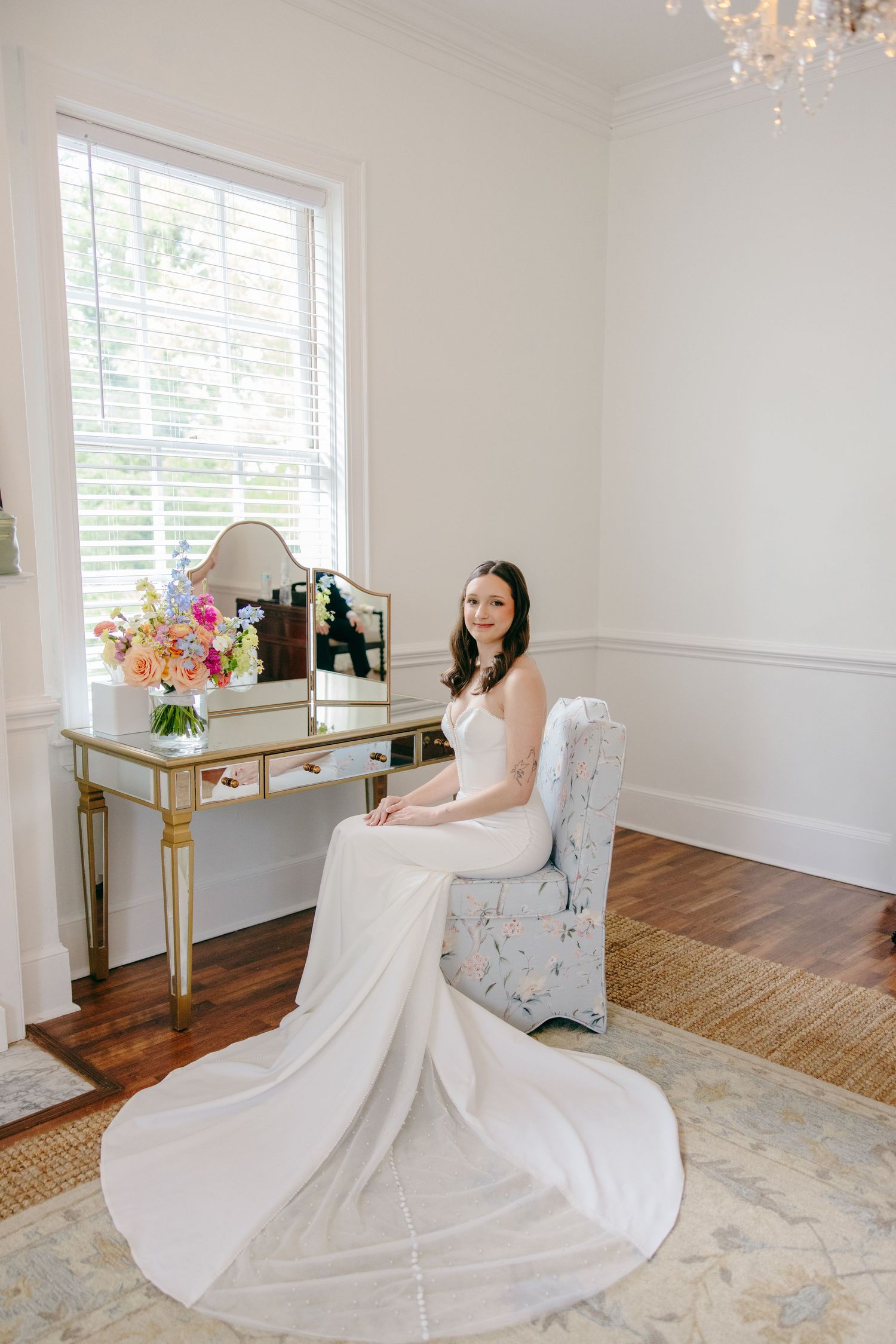 A bride in a flowing white wedding gown sits gracefully in an elegant room with hardwood floors and bright windows.