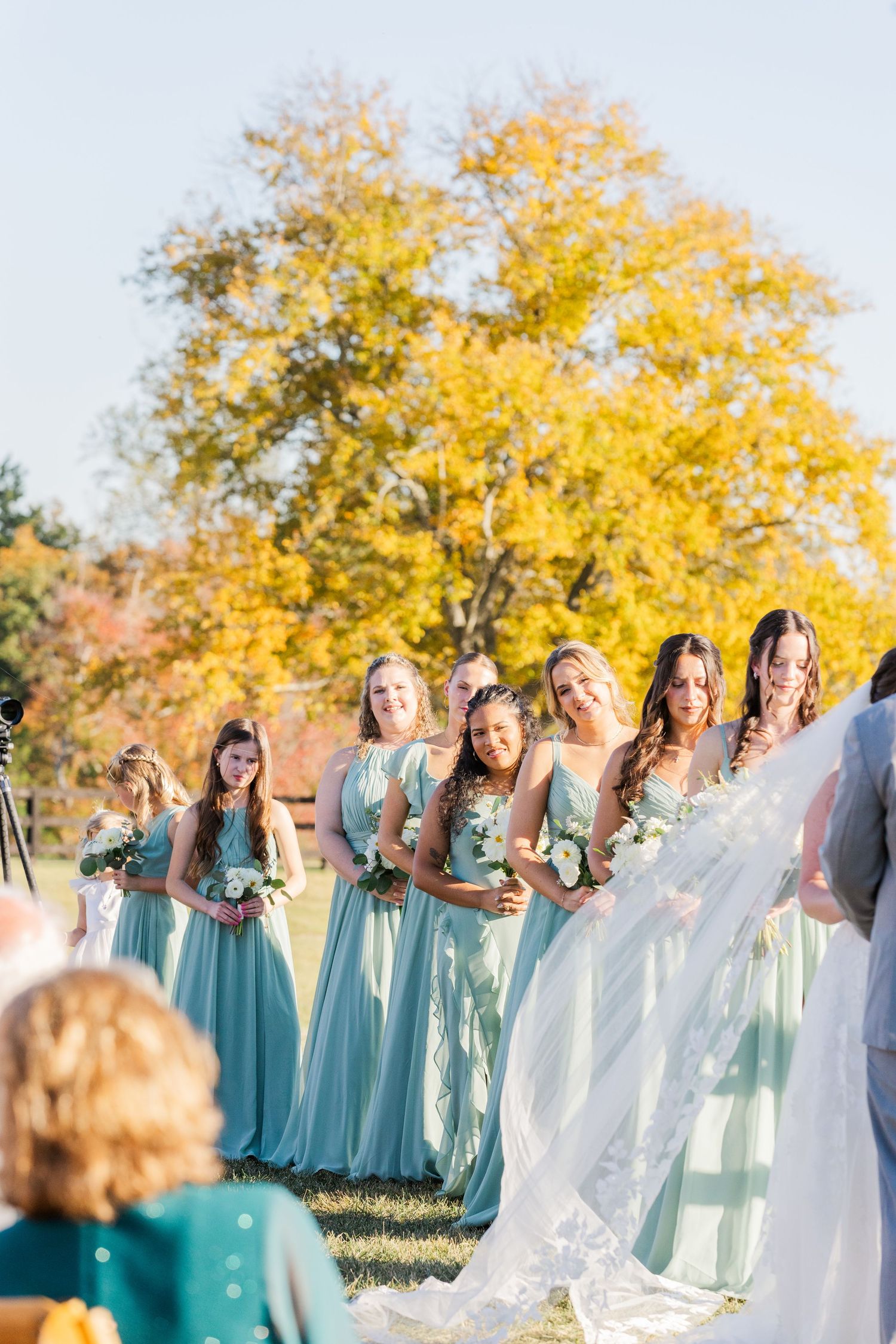 Bridesmaids in turquoise dresses pose together against autumn trees with golden foliage.