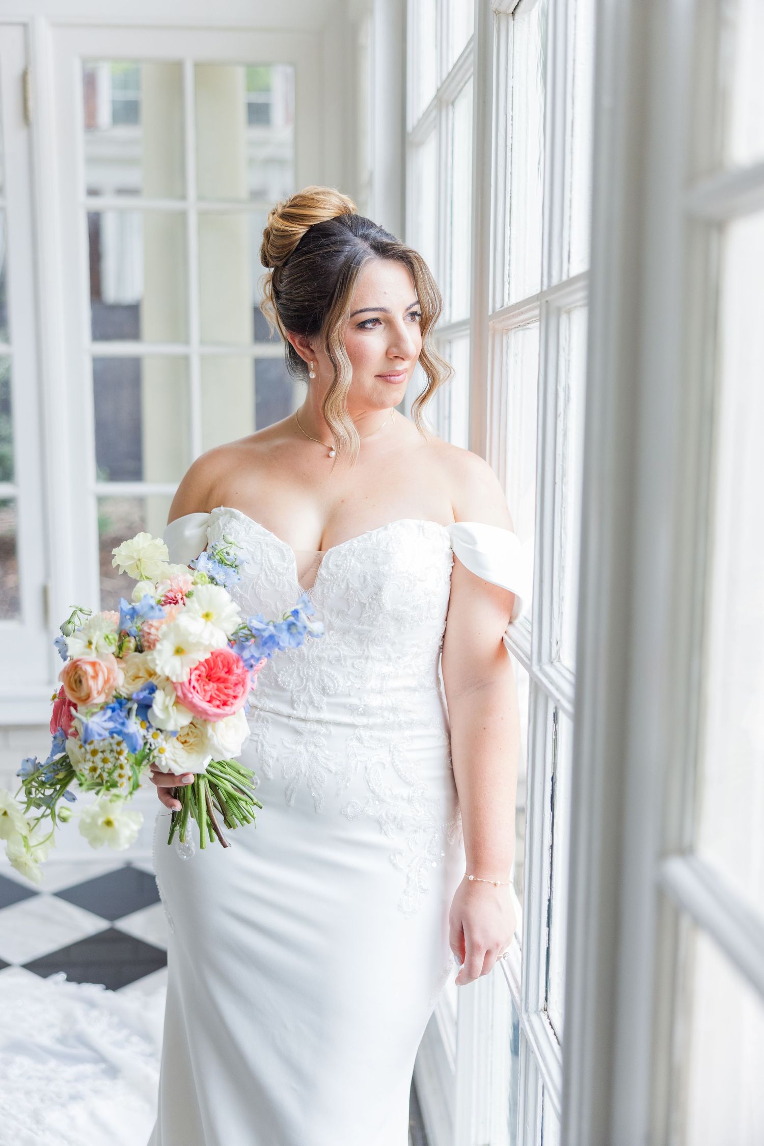 Bride in off-shoulder lace gown holds colorful bouquet by sunlit windows.