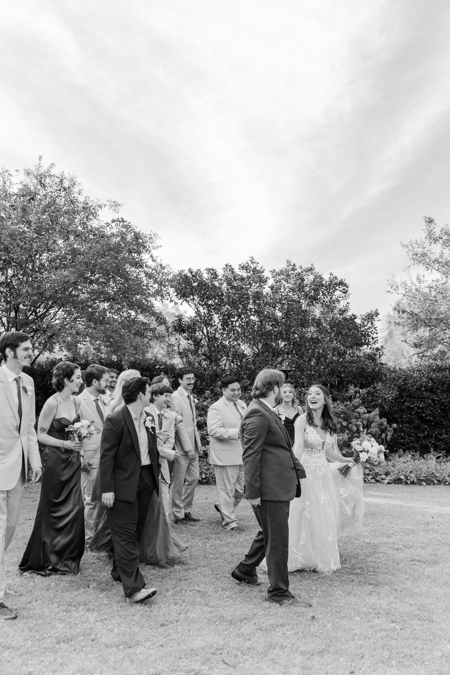 Wedding party walking together outdoors on grassy lawn with trees in background.