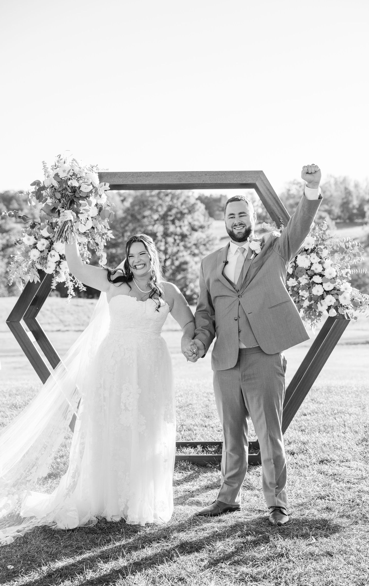 Wedding couple celebrating with joyful poses by geometric wooden arch decorated with white flowers.