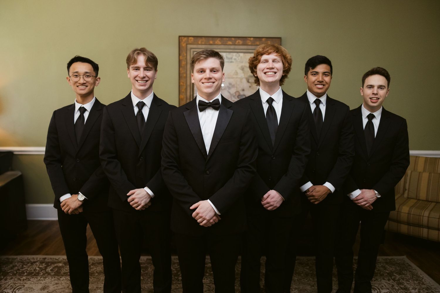 A formal group photo of groomsmen wearing black tuxedos standing in a row at a wedding.