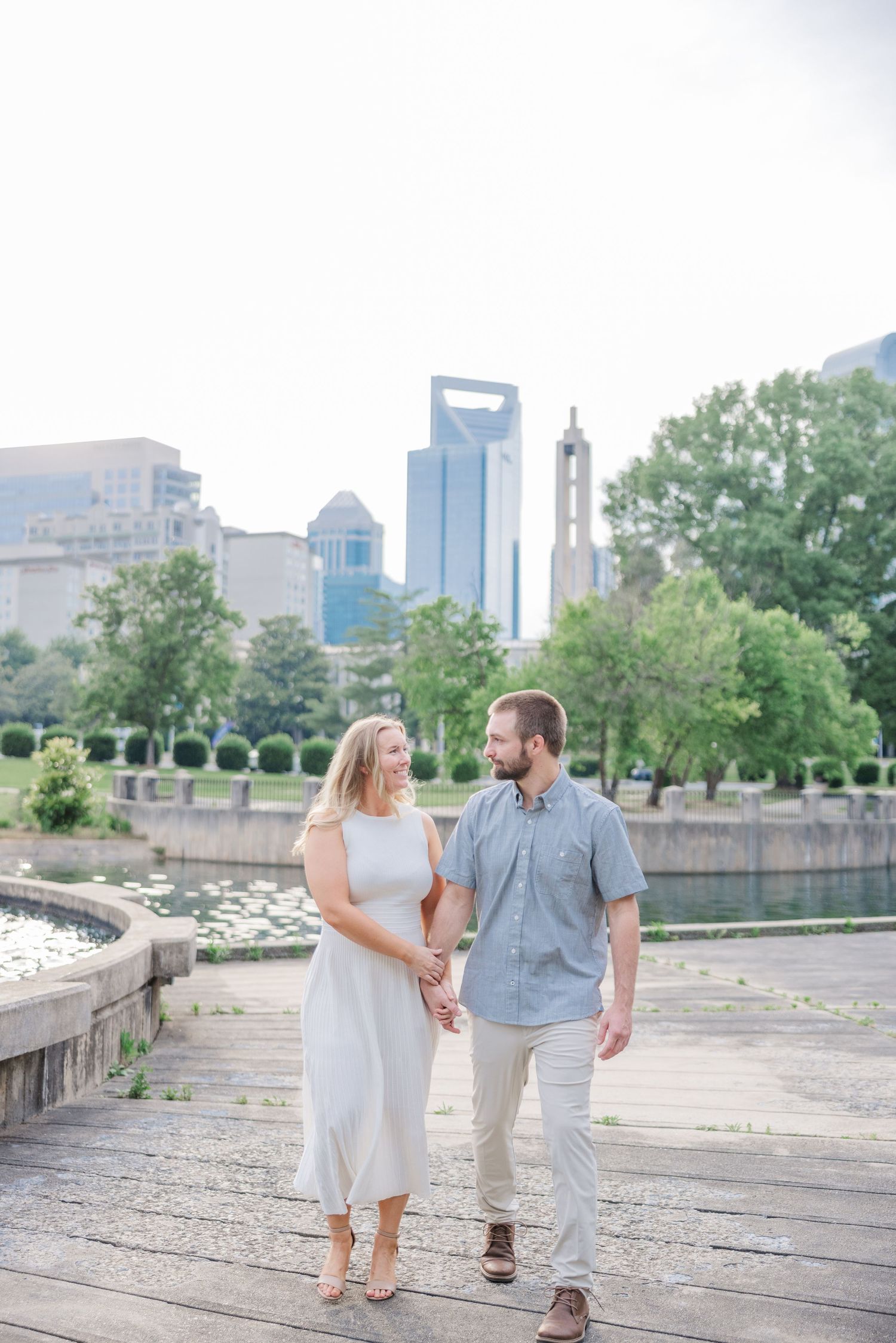 A couple takes a romantic stroll along a waterfront path with urban architecture in the background.