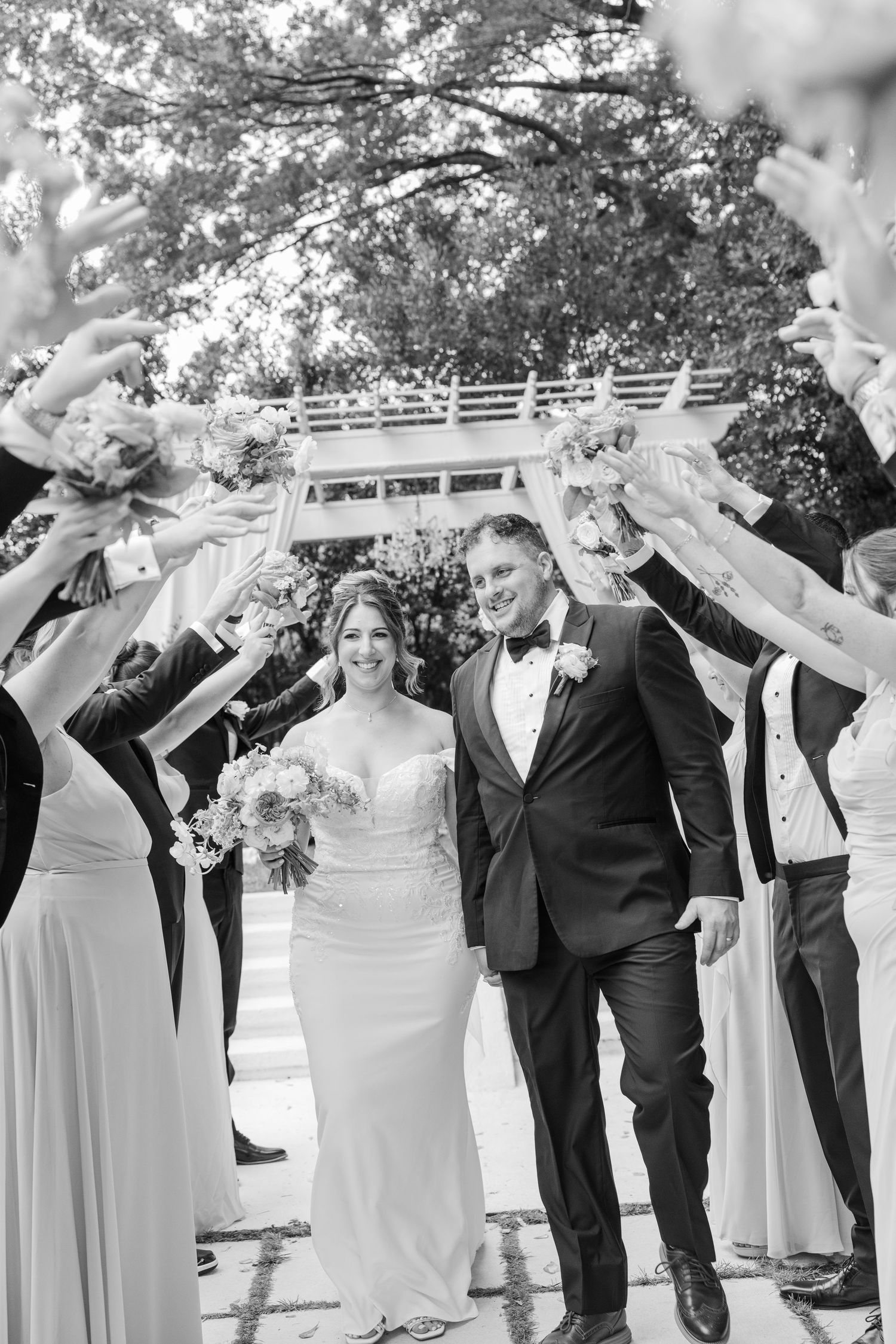 Wedding party in formal attire poses together outdoors with raised arms in celebration.