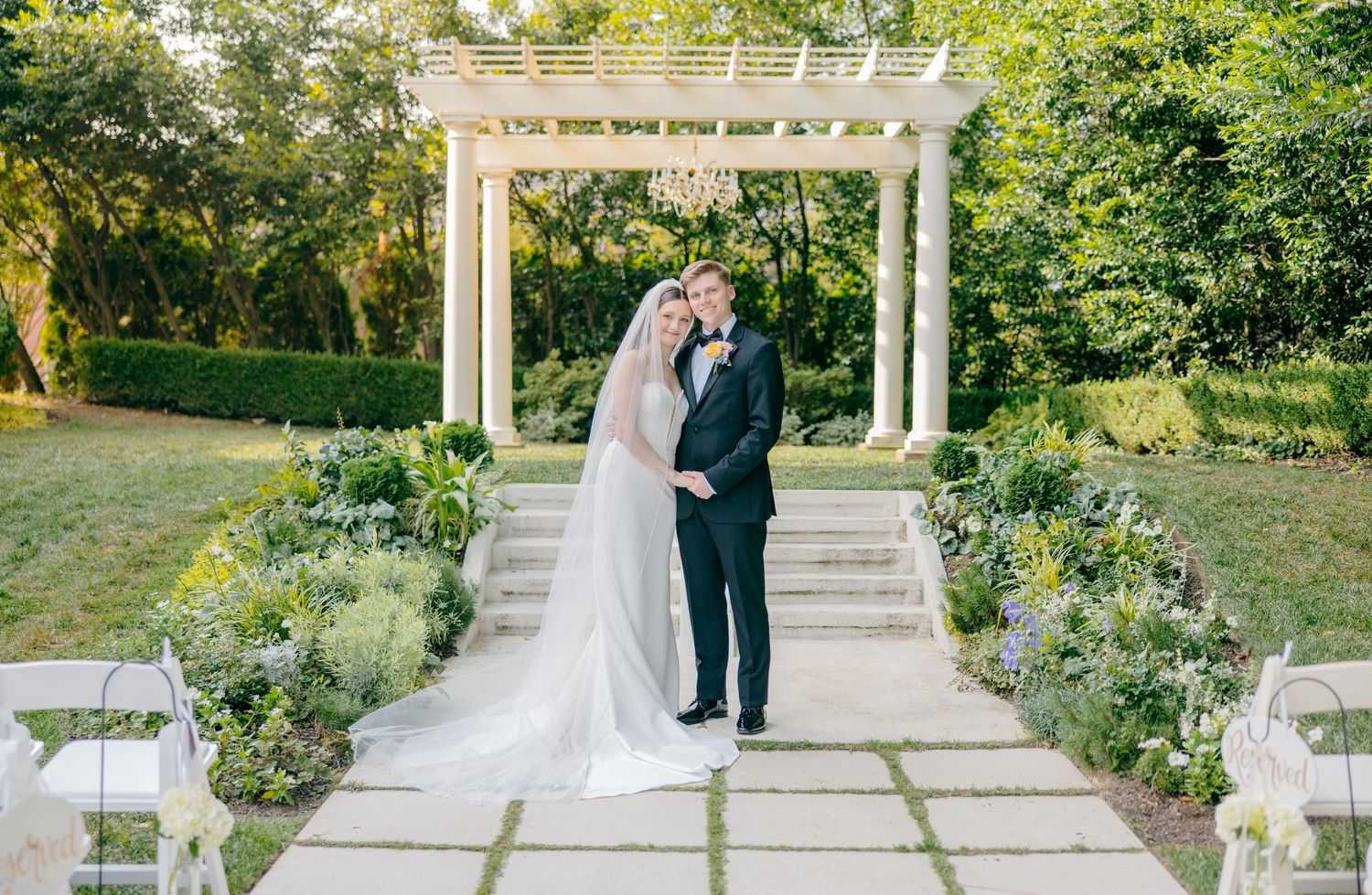 Wedding ceremony setup with white pergola and garden landscaping in outdoor setting.