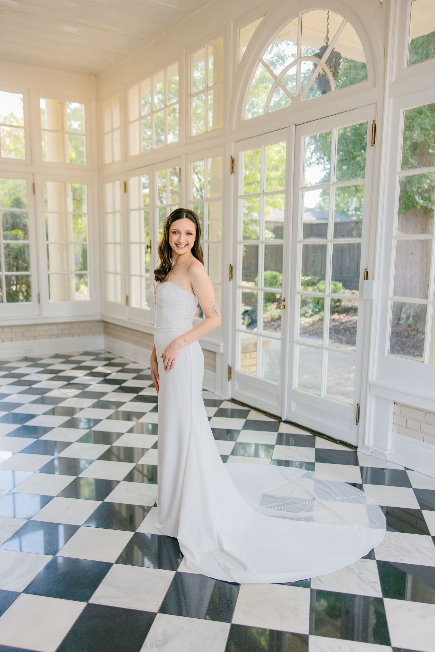 A flowing white wedding dress twirls gracefully across a black and white checkered floor in a sunlit room.