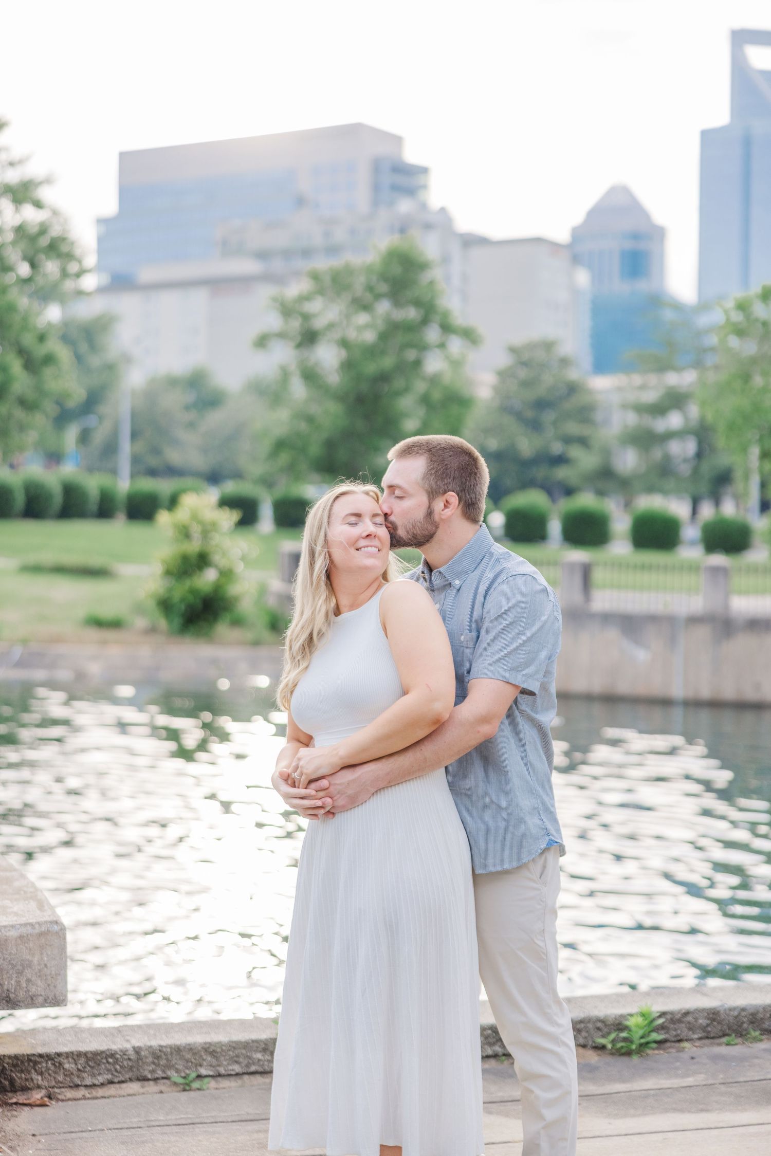 A couple embraces near a reflective pond with city buildings visible in the distance.