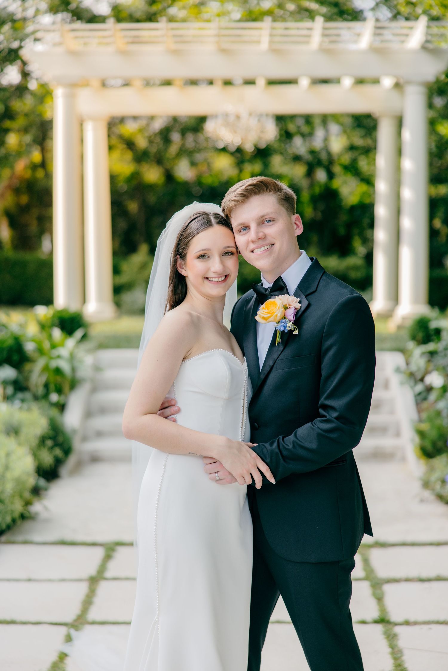 Romantic wedding couple poses under an elegant white garden pergola on a sunny day.